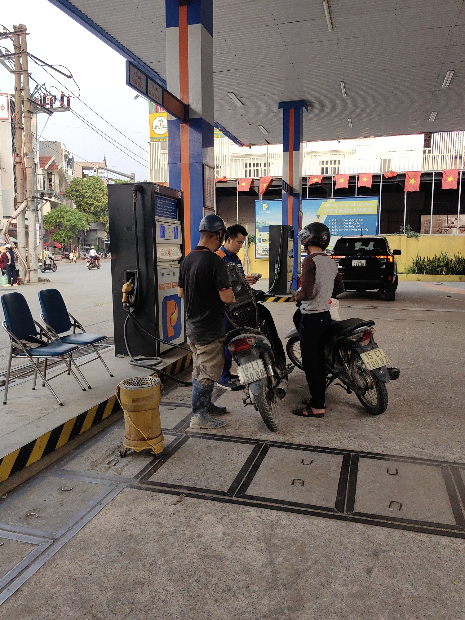 The photograph captures a scene at a Petrolimex gas station in Ha Lam Ward, Vietnam. Three individuals are interacting near a gas pump. One person, wearing a helmet and gray shirt, stands by a motorbike with the license plate "15-C1 100.92". Another individual, wearing a black shirt, helmet, and rubber boots, is standing next to another motorbike with the license plate "18-L1 50.88" and appears to be paying. The third person, dressed in what looks like a gas station uniform, is present near the motorbikes.
The foreground features a yellow, cylindrical trash can with a rope attached to it, and a segment of sidewalk with a black and yellow striped edge. Two blue chairs are positioned to the side. The gas pump itself has a black and orange design with a price display and payment options.
Behind the individuals and motorbikes, a black SUV with the license plate "14A 366.35" is parked. Red and yellow flags hang above the station, possibly Vietnamese national flags. A sign above the pumps advertises "PETROLIMEX" and promotes various types of motor oil. The background reveals buildings, trees, and a street scene with other motorbikes and pedestrians. Utility poles with electrical wires are also visible in the backdrop. The overall setting appears to be a typical urban gas station environment in Vietnam.
