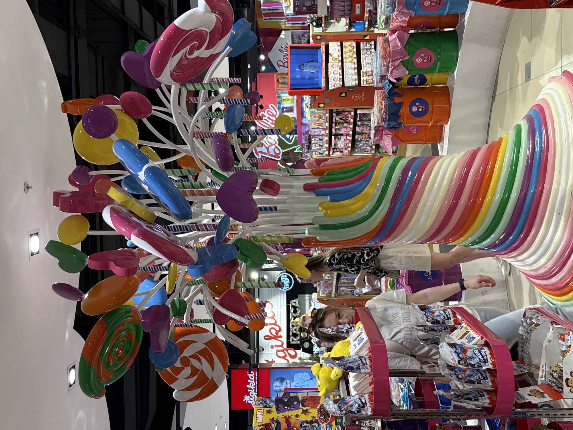 This detailed description pertains to an image captured inside the Central Children's Store (ЦДМ на Лубянке) in Moscow, Russia. The primary subject is a massive, colorful, multi-branched sculpture resembling a candy tree, positioned centrally in the retail space. The sculpture's base is a wide spiral of pastel-colored bands, transitioning into a white central trunk from which numerous white branches extend. These branches support oversized, glossy, artificial candies in various shapes and vibrant colors, including large swirled lollipops (green/orange, orange/white, pink/white), flat disk-shaped candies (yellow, orange, purple), heart-shaped candies (pink, purple, green), and elongated cylindrical candies (blue, pink). Numerous smaller, striped candy canes or sticks are also integrated into the branches. In the foreground, slightly to the left, a pink, multi-tiered display rack holds multiple packages of Kinder brand chocolates, specifically "Kinder Surprise" or similar products, some featuring the text "CHEER WIN" and images of male figures. Behind this rack, several yellow stuffed toys are visible. A woman with light brown hair, wearing a light gray t-shirt with the text "THROW THE RULES" and light blue jeans, stands behind the rack, looking towards the right side of the frame. Her left wrist bears a dark-colored smartwatch, and she appears to be holding one of the Kinder packages. Another woman, with long dark hair, wearing a patterned long-sleeved shirt and light-colored shorts, stands further back on the right, holding a clear plastic bottle. Her gaze is directed towards the left. A third person's head is partially visible behind the foreground woman. The background reveals a brightly lit and spacious children's retail environment. To the left, a red illuminated sign with "MARVEL" branding is visible above shelves stocked with related merchandise. Below this, a red sign with "sigikid GERMAN FOR KIDS" is displayed next to a blue-tinted screen showing a child's face. Further into the store, a large pink sign with the "Barbie" logo marks a section primarily dedicated to Barbie dolls and merchandise, visible on multiple shelves. A red-framed display case showcases a doll. Below the Barbie section, a row of colorful cylindrical barrels (orange, yellow, green, blue) with "Candy Land" logos are arranged on a white platform. These barrels are topped with what appear to be bulk candies or sweets, covered with colorful tissue paper. Overhead, the ceiling is white with embedded rectangular and circular light fixtures, interspersed with black structural elements. A sign with Cyrillic script "ДЕТСКИЙ КАРУСЕЛЬ" (Children's Carousel)