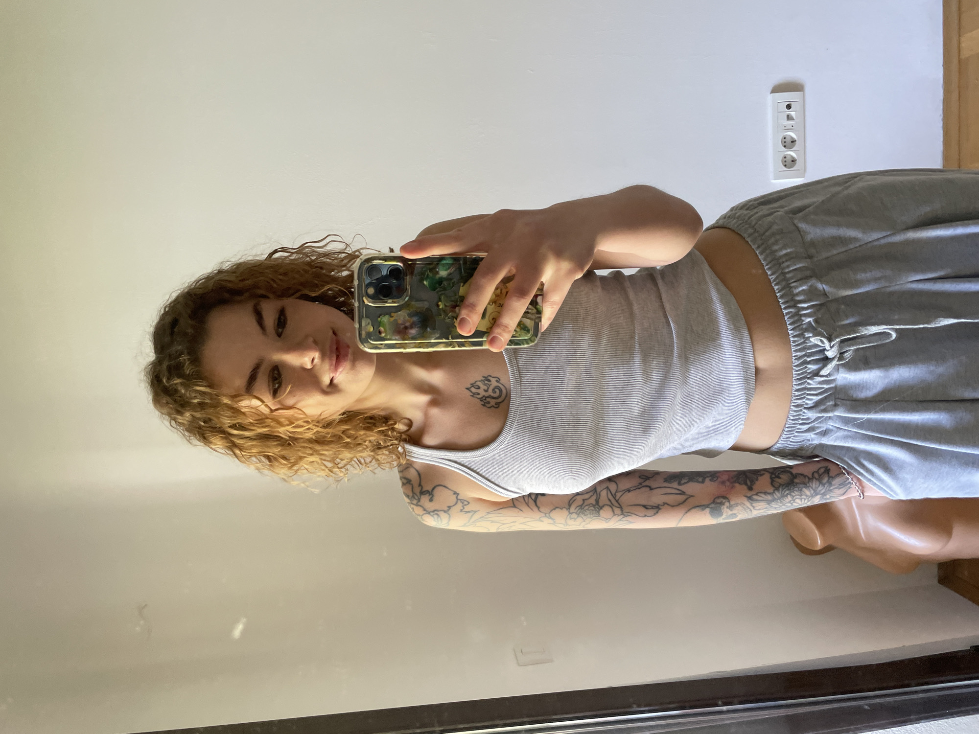 A young woman with curly blonde hair takes a selfie in what appears to be an indoor setting. She is wearing a grey tank top and grey sweatpants. Tattoos are visible on her arm and chest. She holds a phone with a patterned case. Behind her, a wall is visible with a European-style power outlet. Based on the prompt, the location is Boreti, Montenegro, but no information in the picture confirms that.