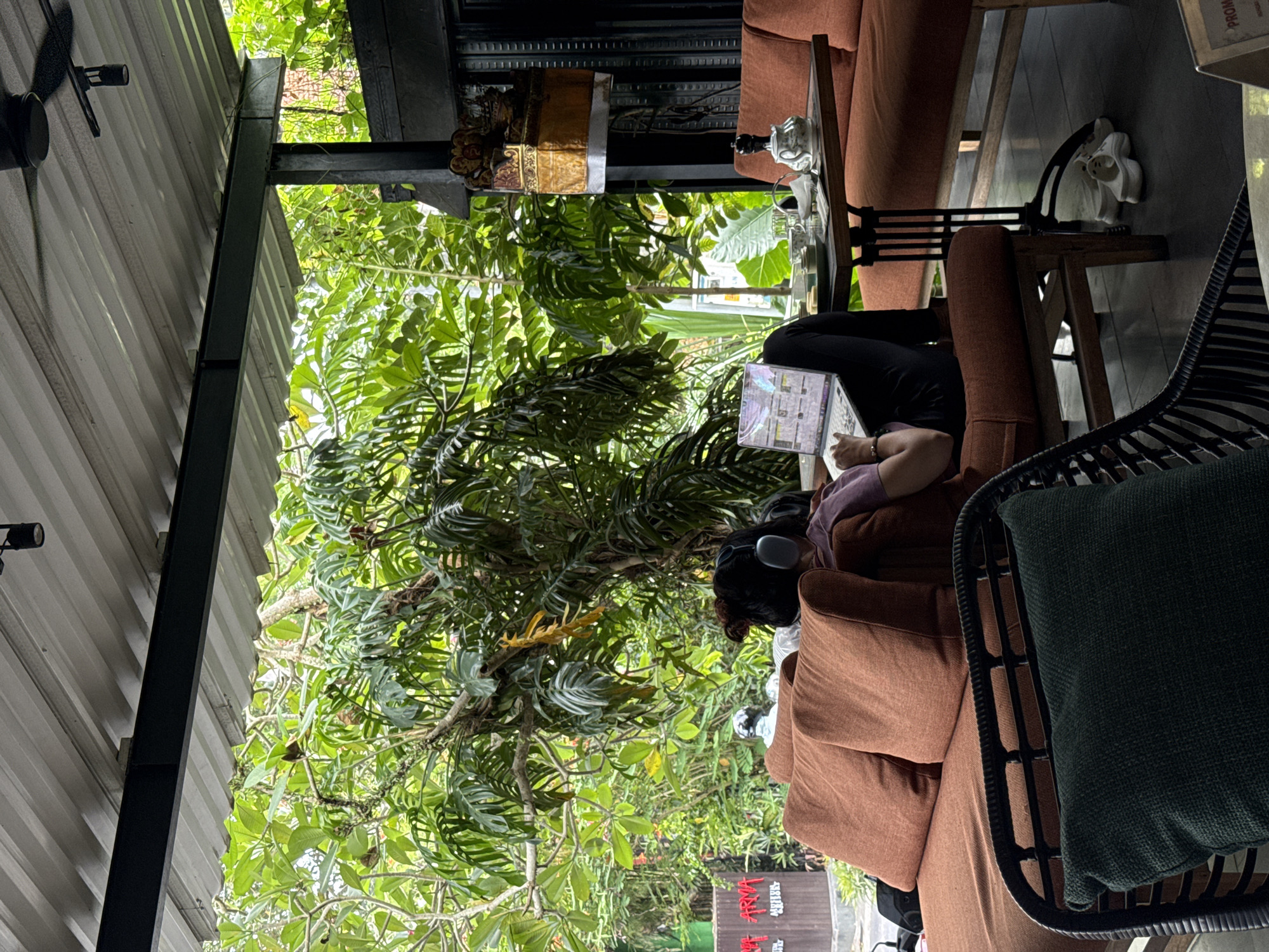 The image shows a person sitting on an orange couch in an outdoor setting, possibly a cafe or restaurant, in the Ubud District, Indonesia. The person is using a laptop. The surrounding environment is lush with green foliage and trees. The seating area is partly covered by a roof structure. A small table with some items is positioned near the person. A wicker chair with a dark green pillow is situated next to the couch. A building structure is visible in the background. A sign with "ARYA" is also visible.