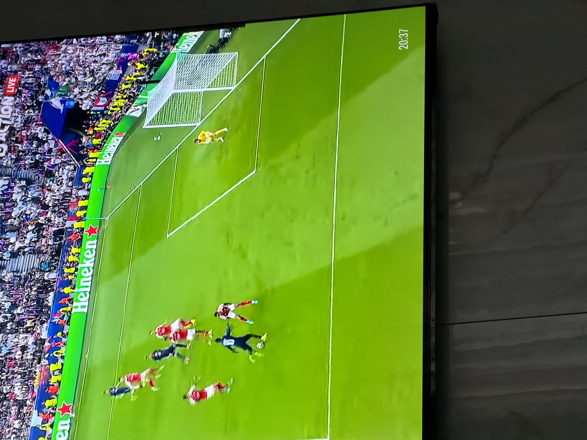 The image shows a television screen displaying a soccer game in progress. The camera is positioned above the field, providing an aerial view of the players, the goal, and a segment of the crowd. A group of players is clustered near the center of the field, while the goalkeeper is positioned near the goal. The time displayed on the screen is 20:37. The location in Asenovgrad, Bulgaria is not apparent.