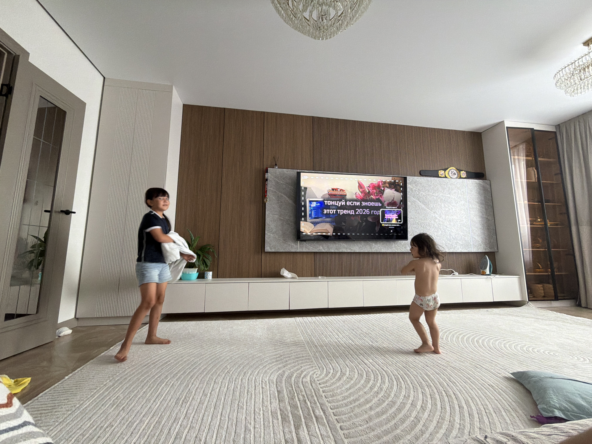 Two children, a boy and a younger girl, are in a brightly lit, modern living room. The boy, wearing a dark blue t-shirt and light denim shorts, stands on a large, textured cream rug, holding a white piece of fabric. The younger girl, wearing patterned underwear, is barefoot and dancing in the center of the rug. Behind them, a long, low white media console spans the wall, topped by a large flat-screen television displaying a video with Russian text. Above the television, a championship belt is displayed on the wall. The room features wood-paneled walls, a large window with grey curtains, and ornate chandeliers on the ceiling. The overall atmosphere is one of playful activity and domestic comfort. The time of day appears to be daytime, judging by the bright natural light filtering into the room. The location is identified as Astana, Kazakhstan. The text on the TV screen is in Russian and reads "танцуй если знаешь этот тренд 2026 год" which translates to "dance if you know this trend 2026 year".
