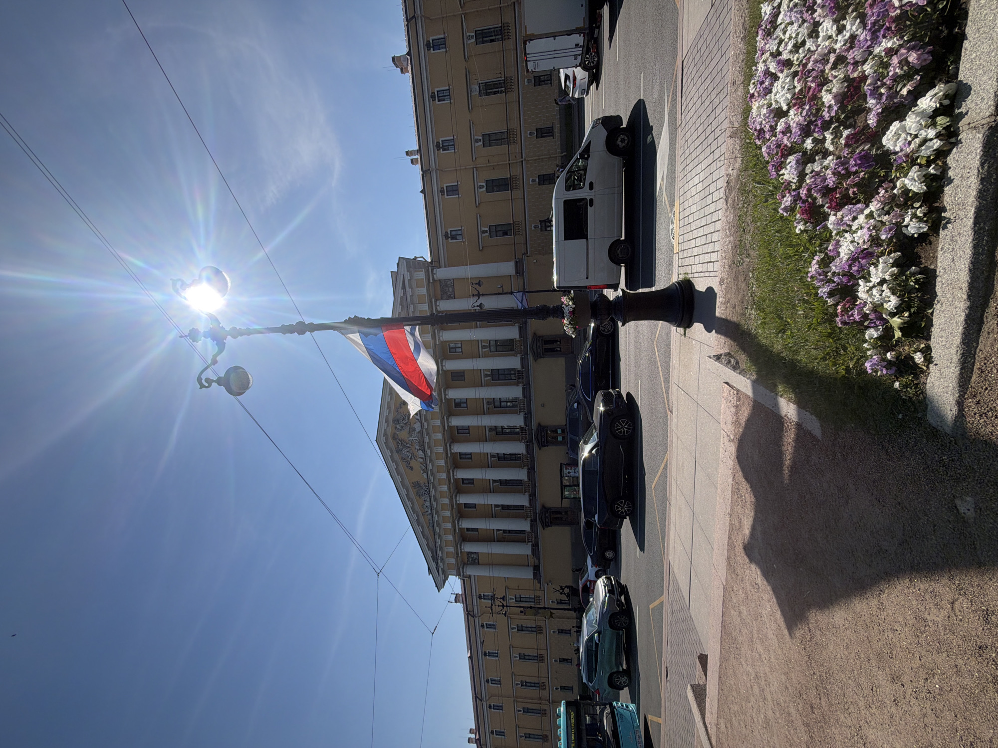 The image shows a sunny day in Saint Petersburg, Russia. In the foreground, a patch of colorful flowers, primarily white, purple, and pink, lines a sidewalk. A shadow of a lamppost is cast on the ground. The lamppost itself is tall and dark, adorned with a flower planter and a Russian flag flying from it. Behind the sidewalk, a street is visible with several cars, including a white van and a dark SUV, driving on it. Beyond the street, a large, classical-style building with yellow walls and white columns dominates the background. The building has a grand facade with multiple windows and decorative elements on its roof. The sky is clear blue with a bright sun, causing lens flare in the image. The scene appears to be a public space, likely a square or a wide street in the historical center of the city.