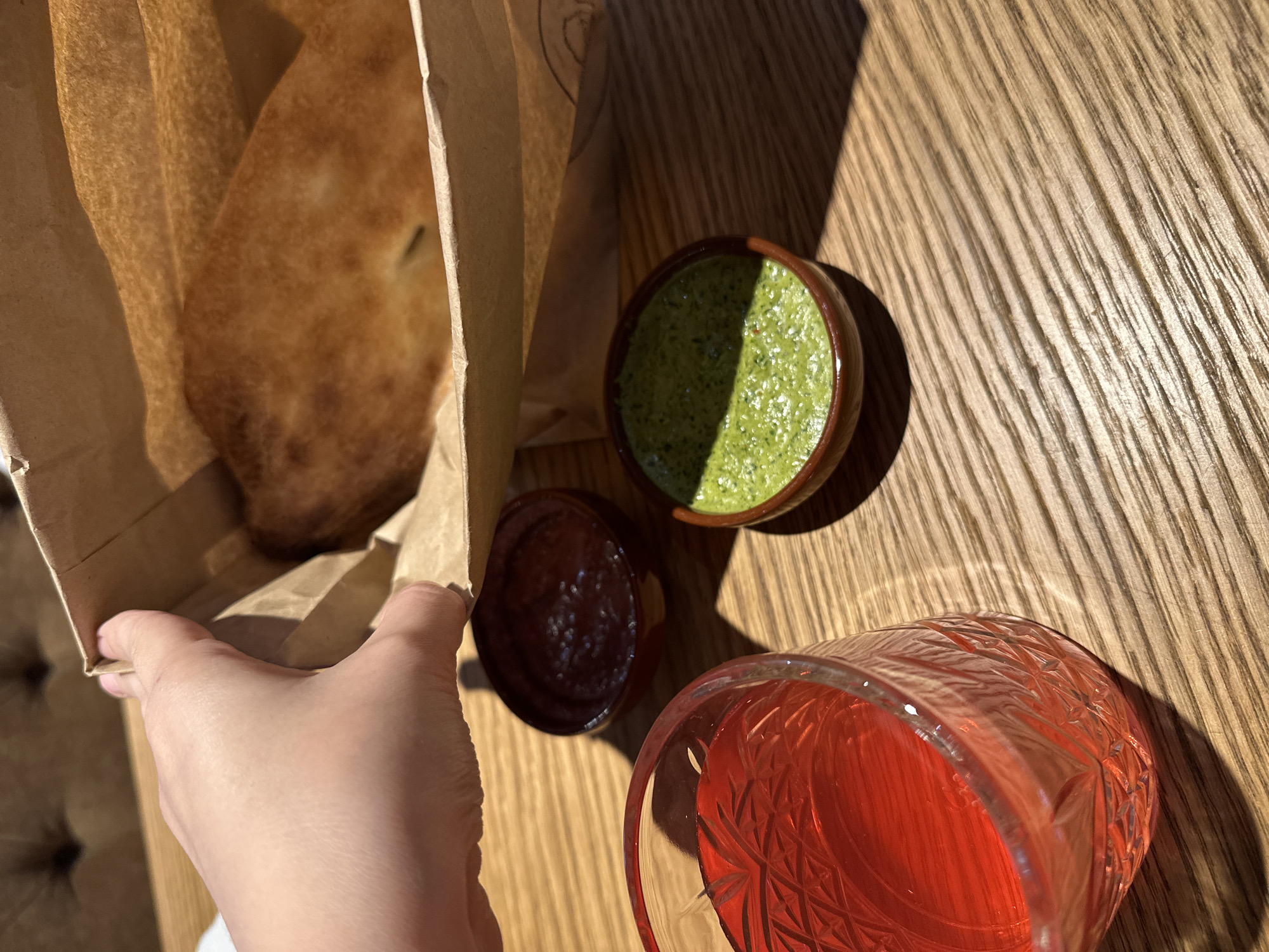 The scene depicts a table setting in what appears to be a restaurant. A hand holds a brown paper bag containing a piece of flatbread. Adjacent to the bag are two small bowls, one containing a green sauce, potentially pesto, and the other a red sauce. A glass of red liquid is positioned near the bowls. The table has a light brown wood grain surface, and the lighting suggests it is daytime. The location is not identifiable as being in Izhevsk, Russia, or any other specific place without additional context.