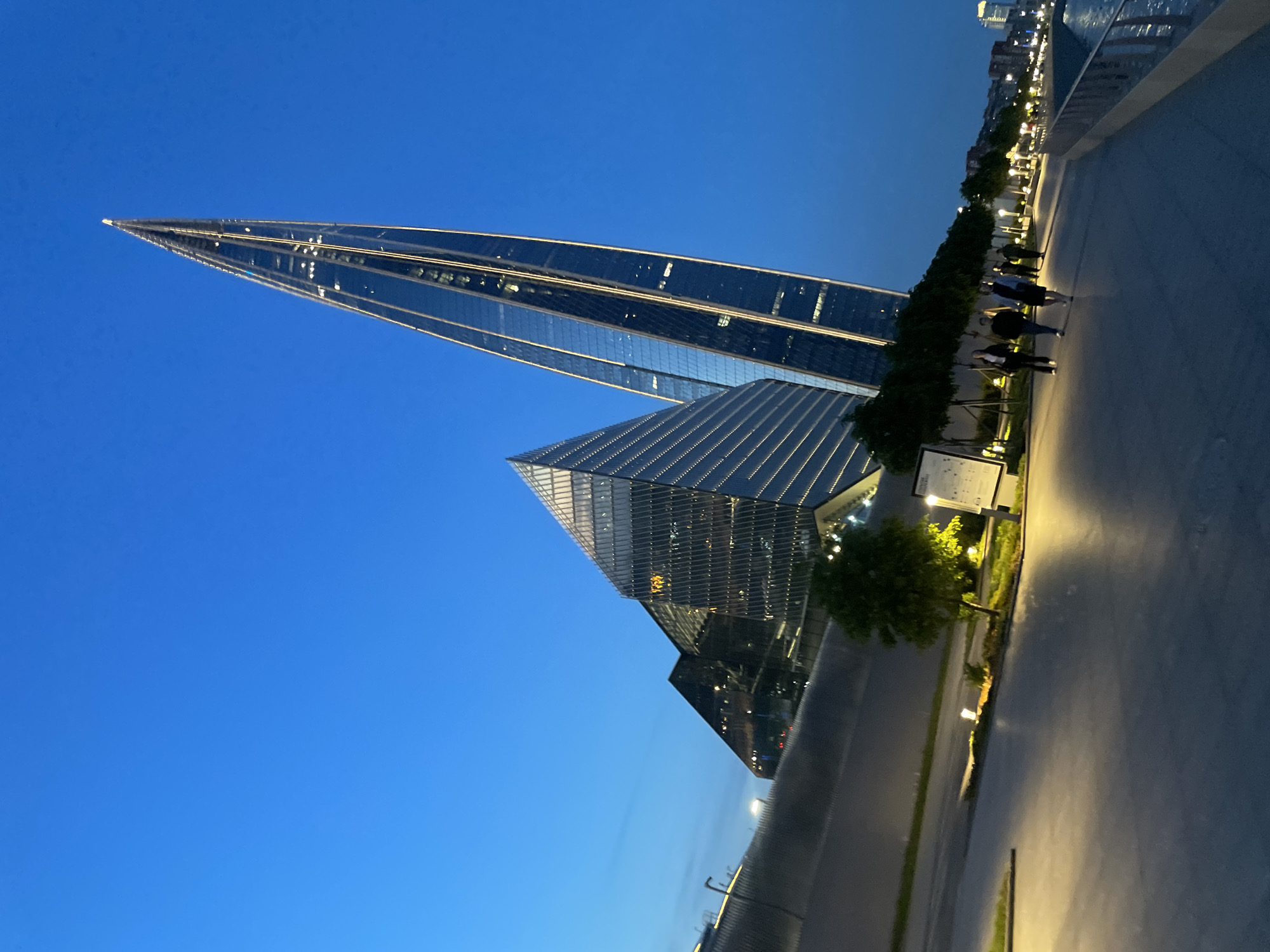 The image depicts the Lakhta Center in Saint Petersburg, Russia, at dusk. The distinctive twisting skyscraper, illuminated by golden lights along its edges, dominates the right side of the frame. To its left, a large, angular, glass-fronted building, also lit, reflects the twilight sky.
In the foreground, a paved promenade runs along a waterfront, with people walking and silhouetted against the light. Trees are planted along the edge of the promenade, and streetlights cast warm pools of illumination on the path. The water of the bay is visible on the right, with a railing separating the promenade from the water. In the background, across the water, city buildings are visible with lights on. The sky is a deep, clear blue, transitioning to a lighter shade near the horizon.
