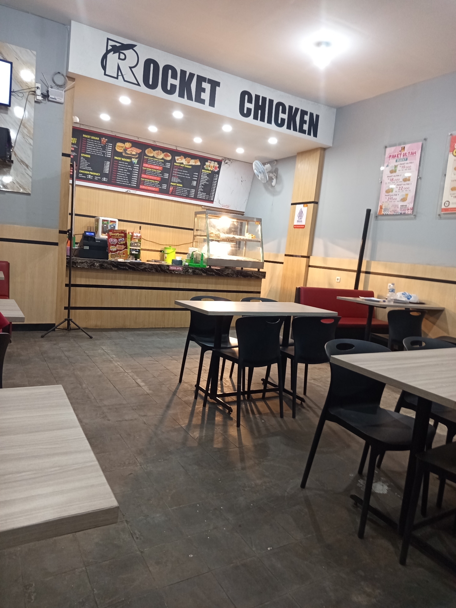 The image shows the interior of a "Rocket Chicken" fast-food restaurant, likely located in Prajekan Lor, Indonesia. The restaurant features a counter area with a cash register, a display case for food, and a menu board above. The dining area is furnished with tables and chairs, some of which are visible in the foreground and midground. The walls are decorated with posters and signage, including a large "ROCKET CHICKEN" logo. The floor is tiled. The overall impression is of a typical fast-food establishment.