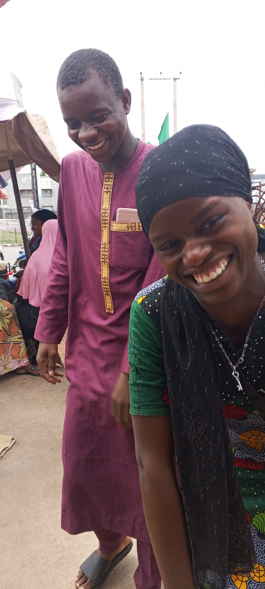 In Maiburiji, Nigeria, a man and a woman are captured on an outdoor street. The man, dressed in a maroon tunic with gold trim and dark sandals, is smiling. The woman, in the foreground, wears a black headscarf and a brightly patterned green and blue top with a silver necklace; she is also smiling. In the background, other people and objects such as awnings and distant vertical structures, are visible.