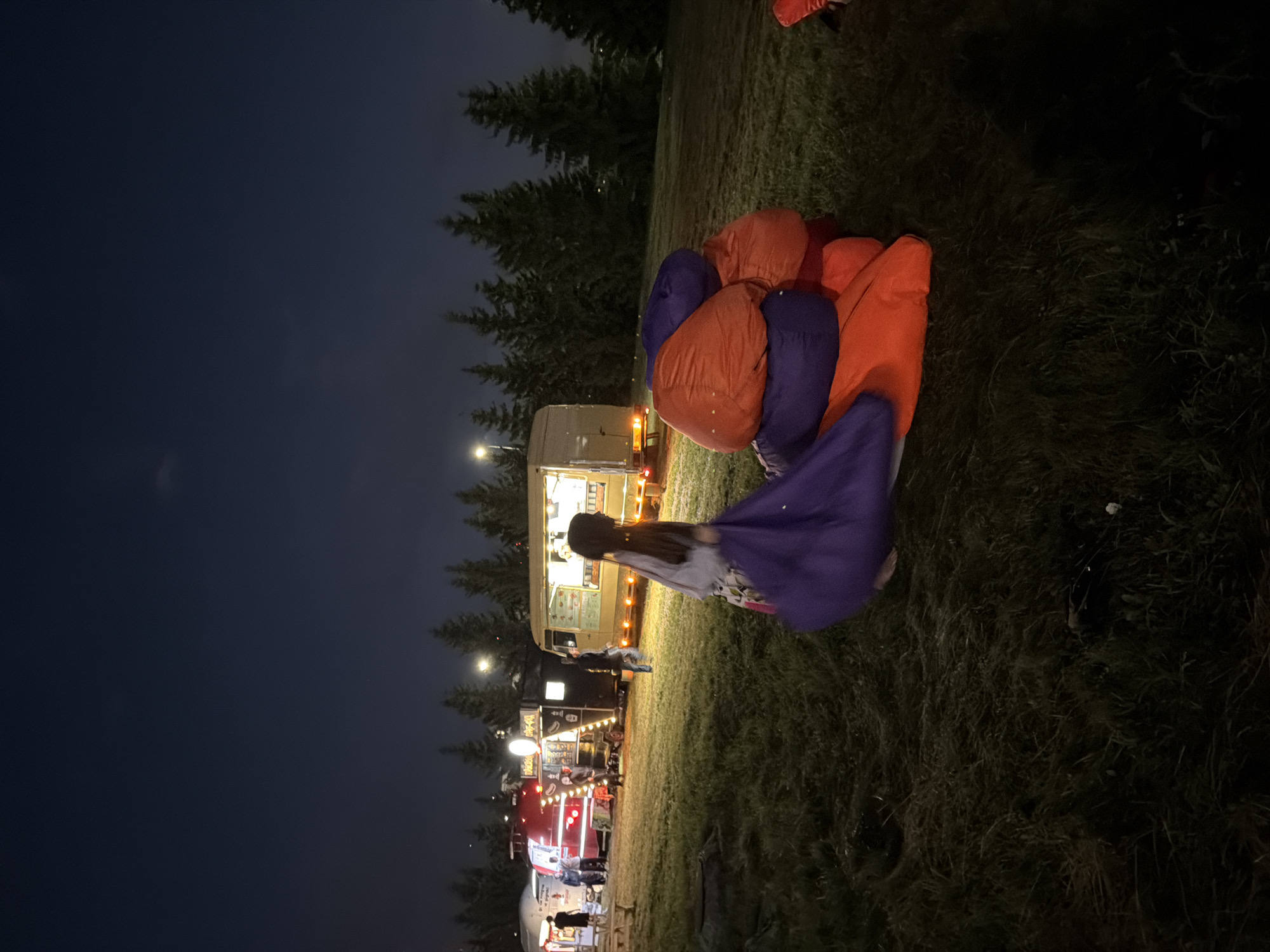 In an outdoor grassy area at dusk, a person wearing a purple and white garment stands beside a pile of large, stacked beanbag chairs in orange and purple. In the background, several food trucks are parked in a row, illuminated by their lights. A line of evergreen trees forms a backdrop against the dark night sky. Several people are visible near the food trucks, some appearing to be customers or employees. The scene suggests a nighttime outdoor event or gathering.