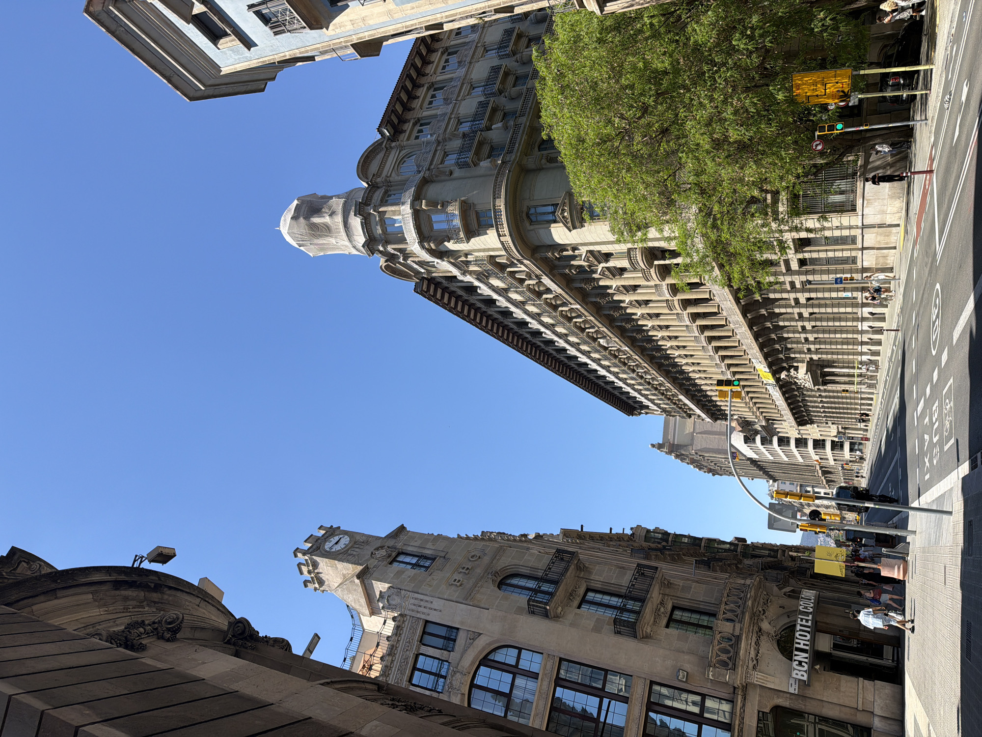 The image depicts a street scene in Barcelona, Spain, viewed from a low angle, looking up at the buildings. The sky is clear and blue. The buildings are primarily constructed from stone and feature various architectural details such as windows, balconies, and decorative elements. One of the buildings is covered in scaffolding and protective netting. A street runs along the right side of the image, with a crosswalk, pedestrian traffic, street signs, and a traffic light. "BCN HOTEL COLON" is seen on one of the buildings. The composition includes trees, further signs, and street lights.