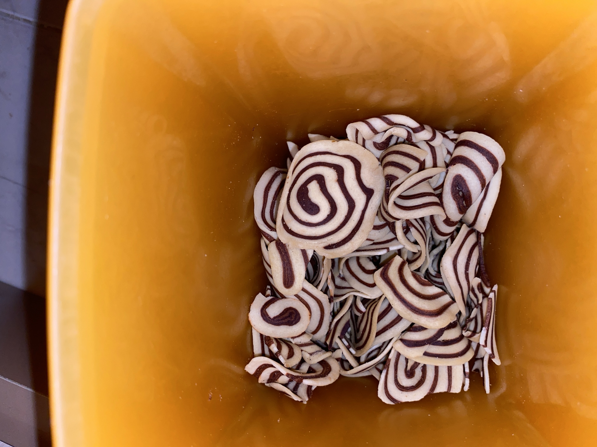 A top-down shot reveals a bright orange container filled with thin, swirled cookies. The cookies are predominantly beige and brown, with the brown forming concentric circles within the beige. Some cookies are whole, showing a spiral pattern, while others are broken into fragments, revealing layers of the beige and brown swirl. The background outside the container is dark and indistinct. The location and city of Lhokseumawe, Indonesia are not discernible from the visual information.