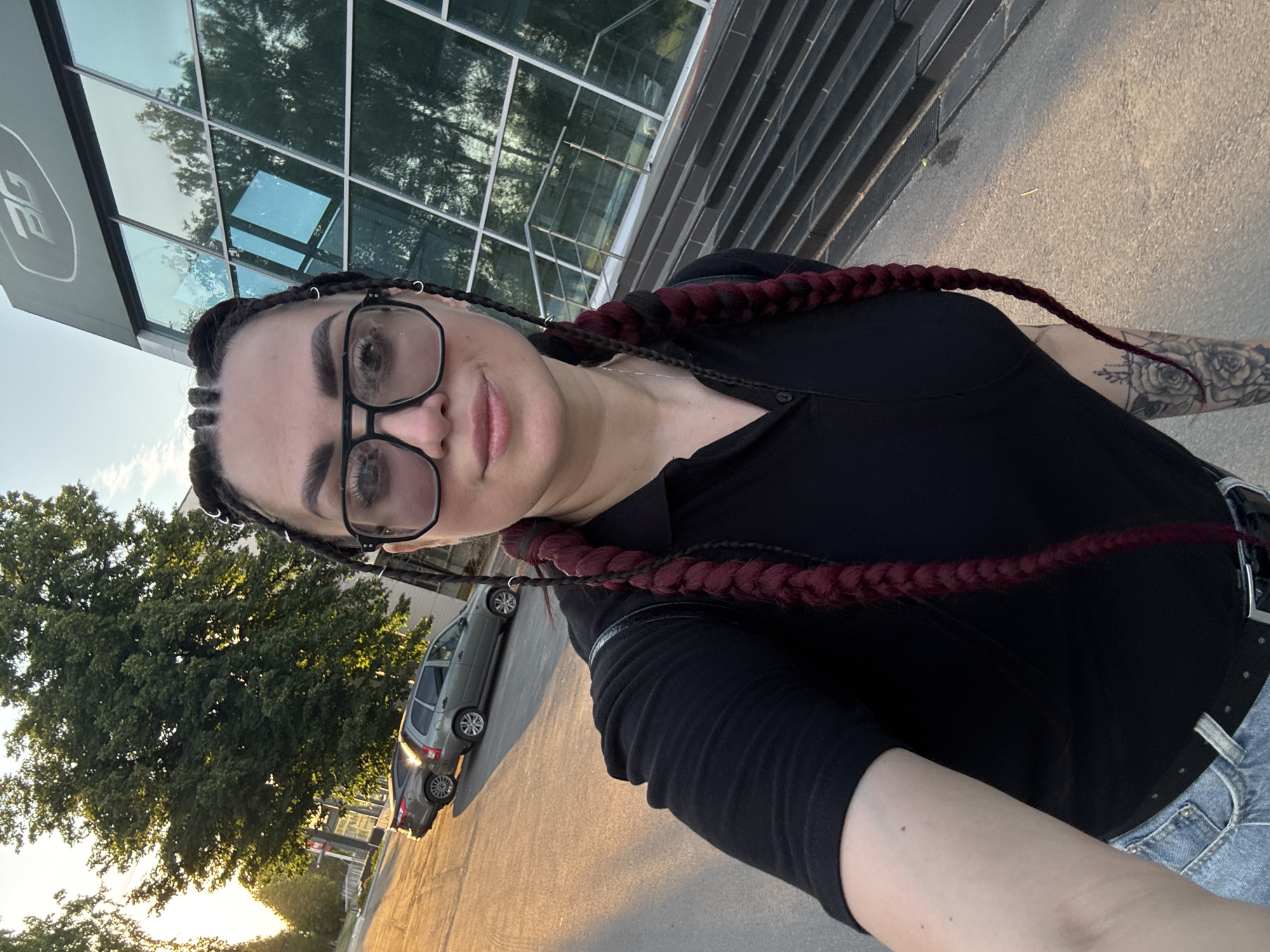 In an outdoor, urban setting, a woman with dark hair styled into braids and wearing large-framed glasses takes a selfie. She is dressed in a black polo shirt and denim jeans. Her left arm is visible, showing a tattoo of roses on her forearm. Behind her, a modern building with reflective glass windows and a staircase is visible. The sun is setting, casting a warm glow on the scene and reflecting off two parked cars. Trees are present in the background, with one large tree prominently featured to the left. The ground is paved, and the sky is clear.