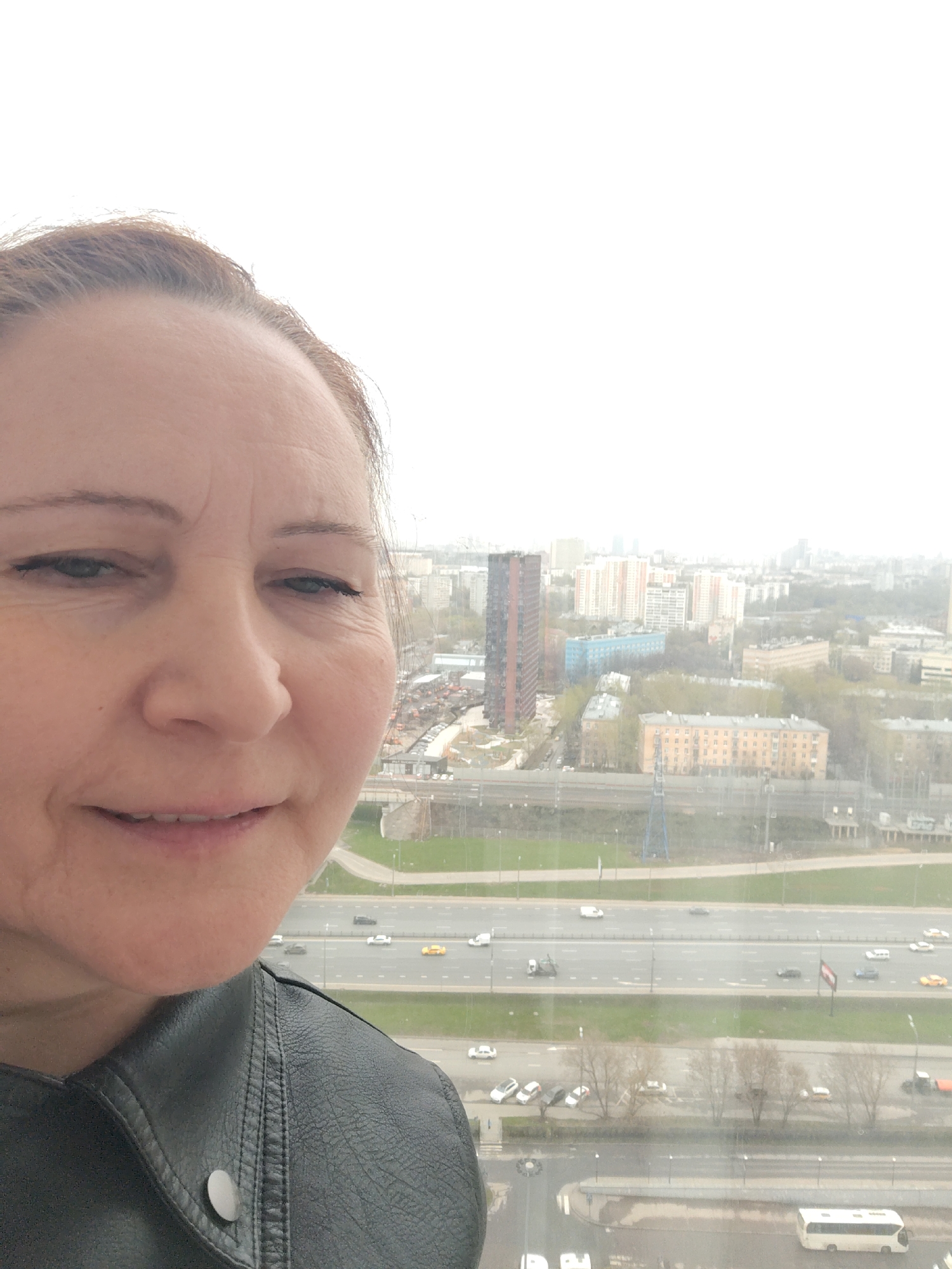A close-up selfie captures a woman in the foreground against a vast urban backdrop, likely taken from a high vantage point overlooking Moscow, Russia. The woman, appearing middle-aged with light skin and light brown hair pulled back, occupies the left side of the frame. She wears a black leather jacket, with a silver snap visible on its lapel. Her expression is a slight, closed-mouth smile, and her right eye appears somewhat squinted, with visible wrinkles around her eyes and on her forehead.
The background reveals an expansive cityscape under an overcast, hazy sky, indicating a cloudy daytime. A multi-lane highway stretches horizontally across the middle distance, busy with various cars, trucks, and a white bus. Beyond the highway, numerous residential buildings of different architectural styles and heights populate the city, including a prominent tall, dark-colored building on the far left and a peach-colored apartment building closer to the highway. Sparse trees, possibly bare due to the season, are scattered throughout the scene. There is no visible text in the image.