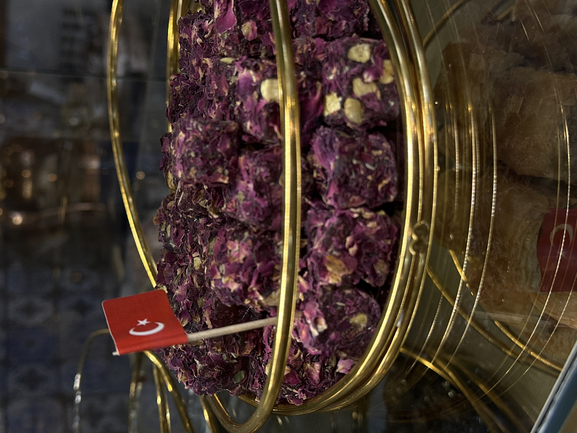 The image presents a close-up view of a display featuring Turkish Delight, adorned with a small Turkish flag, within a glass case likely located in North Kuta, Indonesia. The focus is on the Turkish Delight pieces coated in what appears to be dried rose petals, stacked within a circular, multi-tiered golden metal stand. The stand itself consists of several concentric rings. Another variety of Turkish Delight, possibly coated in nuts, is visible in the background within the same glass case. The lighting is diffused, creating a soft glow on the golden stand. The background is blurry.