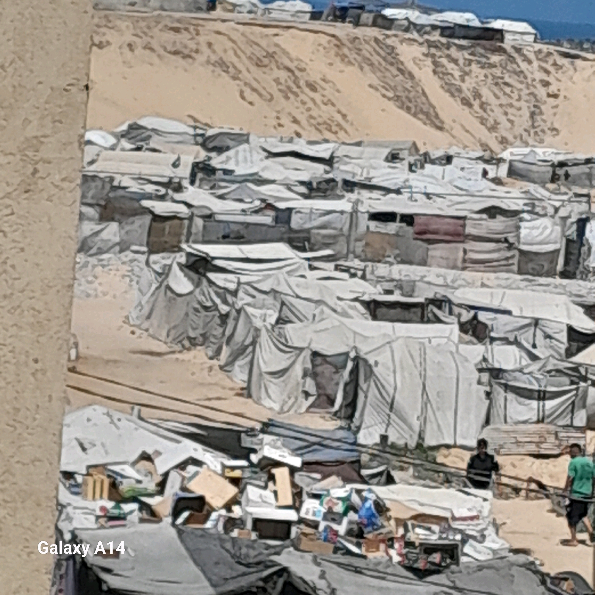 The image depicts a refugee camp in Khan Yunis, Palestinian Territories. The scene is characterized by a densely packed settlement of tents and makeshift shelters, predominantly constructed from white fabric stretched over frames, clustered together on a sandy, uneven terrain. In the background, a steep, sandy incline is visible, dotted with more structures and a body of water at the very top.
In the foreground, a pile of debris, including cardboard boxes, plastic bags, and various discarded items, is scattered on the ground, partially covered by tarpaulins. To the right of the debris, two individuals are present. One person, wearing a dark shirt and trousers, is standing near a wooden fence. Another person, wearing a green shirt and black shorts, is walking away from the viewer. The overall environment appears arid and dusty. The "Galaxy A14" watermark is visible in the lower left corner of the image.
