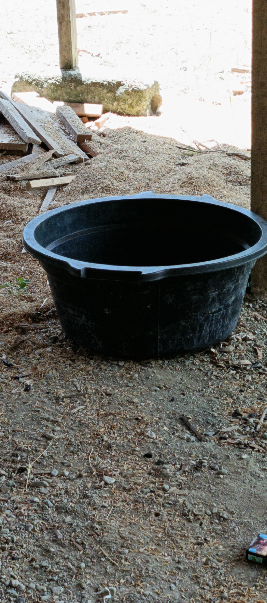 An outdoor scene captures a large, black, plastic tub situated on the ground. The ground appears to be composed of dirt and small pebbles, interspersed with a significant amount of light-colored, granular material, possibly straw or wood shavings. In the background, a wooden post stands vertically, attached to a larger, rough-hewn stone structure. Several pieces of weathered wood are stacked nearby, leaning against the stone. Sunlight illuminates the scene, creating bright areas and shadows, suggesting it is daytime. The environment appears to be rustic, possibly a farm or a workshop area. The location is identified as Meunasahmesjid, Indonesia.