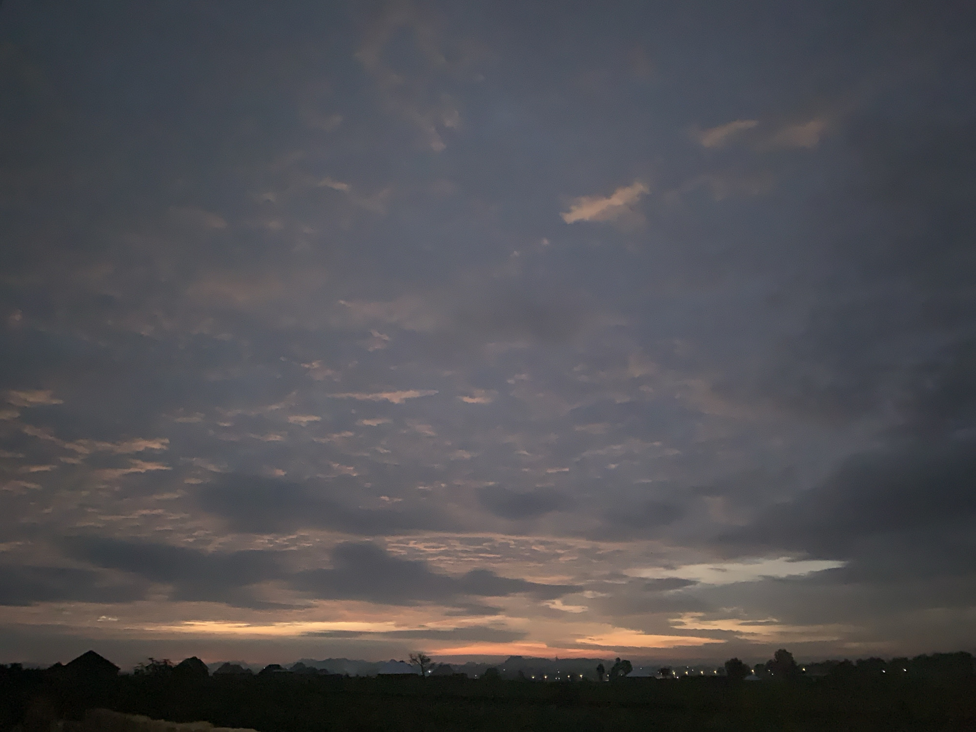 The image depicts a twilight scene over the city of Kuru Karama, Nigeria. The sky is filled with a mix of dark grey and lighter, pinkish-orange clouds, suggesting the last moments of sunset or the early approach of dawn. Below the sky, a dark silhouette of the landscape is visible. This includes several buildings with distinct rooflines, some appearing to be traditional structures with conical or peaked roofs, and others more modern. Trees are scattered across the horizon, also in silhouette. In the distance, faint lights suggest the presence of more buildings or streetlights within the city. The foreground is a dark, indistinct area that appears to be open land. There are no people or animals visible in the frame, and no overt actions or interactions are taking place. The environment is characterized by the darkening sky and the silhouetted features of the urban and natural landscape.