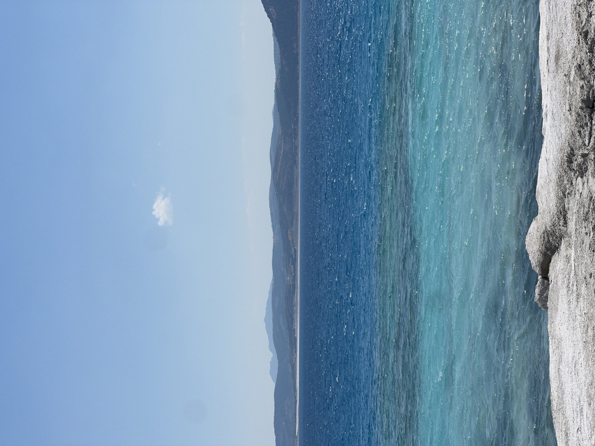 A vertical, eye-level shot captures a vast, shimmering lake in Yeşilova, Turkey. The water transitions from a deep, rich blue in the distance to a lighter, turquoise hue closer to the shore. Gentle waves ripple across the surface, reflecting the bright sunlight. In the foreground, the edge of the lake is lined with a rocky, sandy shore, with a few large, pale rocks visible. The distant horizon is defined by rolling, undulating hills and mountains, partially obscured by a light haze. The sky above is a clear, pale blue, with a single, small, wispy white cloud floating near the top of the frame. The scene is tranquil and natural, with no human presence evident.