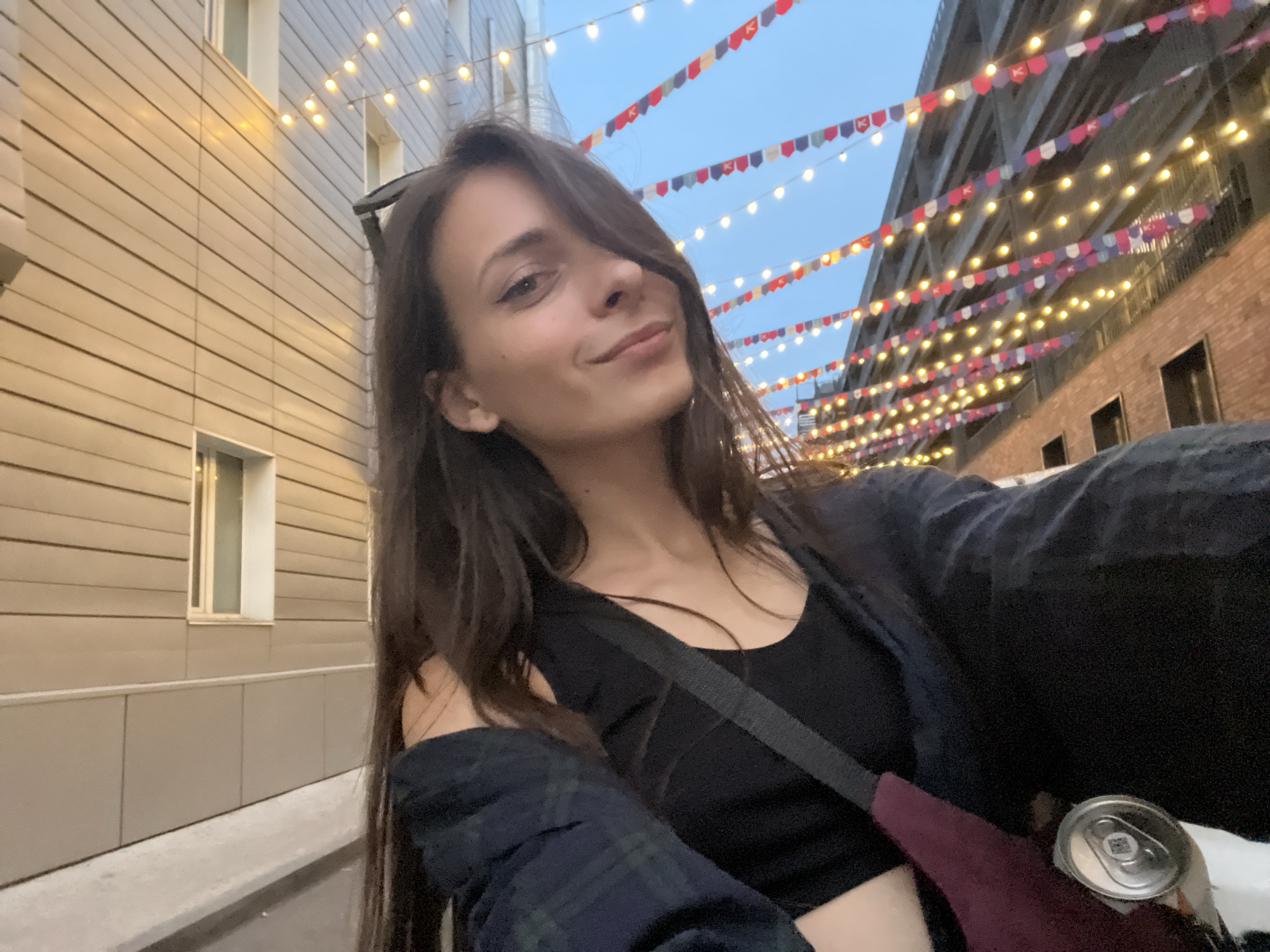 A young woman with long, dark hair is taking a selfie outdoors at dusk. She is wearing a black tank top and a dark plaid shirt, draped off one shoulder. A burgundy bag with a can of soda is visible on her hip. She is standing in what appears to be an alleyway or street decorated with strings of lights and small flags, suggesting a festive atmosphere. The buildings on either side of the alley are modern with horizontal siding on one side and brick with dark-paned windows on the other. The sky is a deep blue, indicating twilight. There is no specific indication of the location within Krasnoyarsk, Russia.