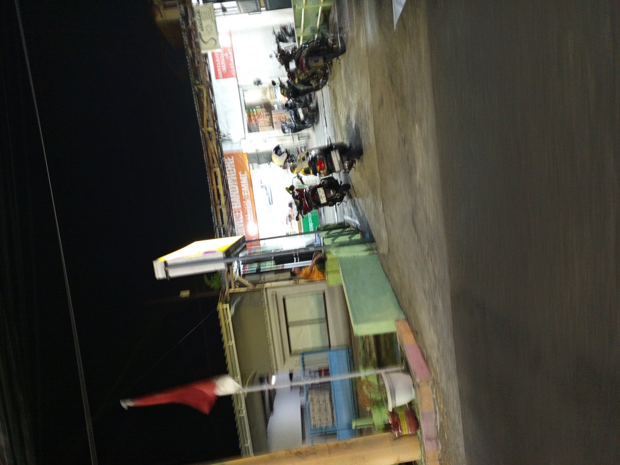 The image depicts an outdoor scene at night in Calukan RT 1 RW 5 Keboansikep, Indonesia. A red and white flag, likely the Indonesian flag, is visible on the left side of the frame, fluttering from a pole. In the background, a building with a yellow illuminated sign that reads "SERVICE HANDPHONE" is present. Several motorcycles are parked in front of the building. A man wearing an orange shirt is seated on a green ledge to the right of the building, facing away from the camera. The ground appears to be paved. The overall impression is that of a street-side establishment open at night.
