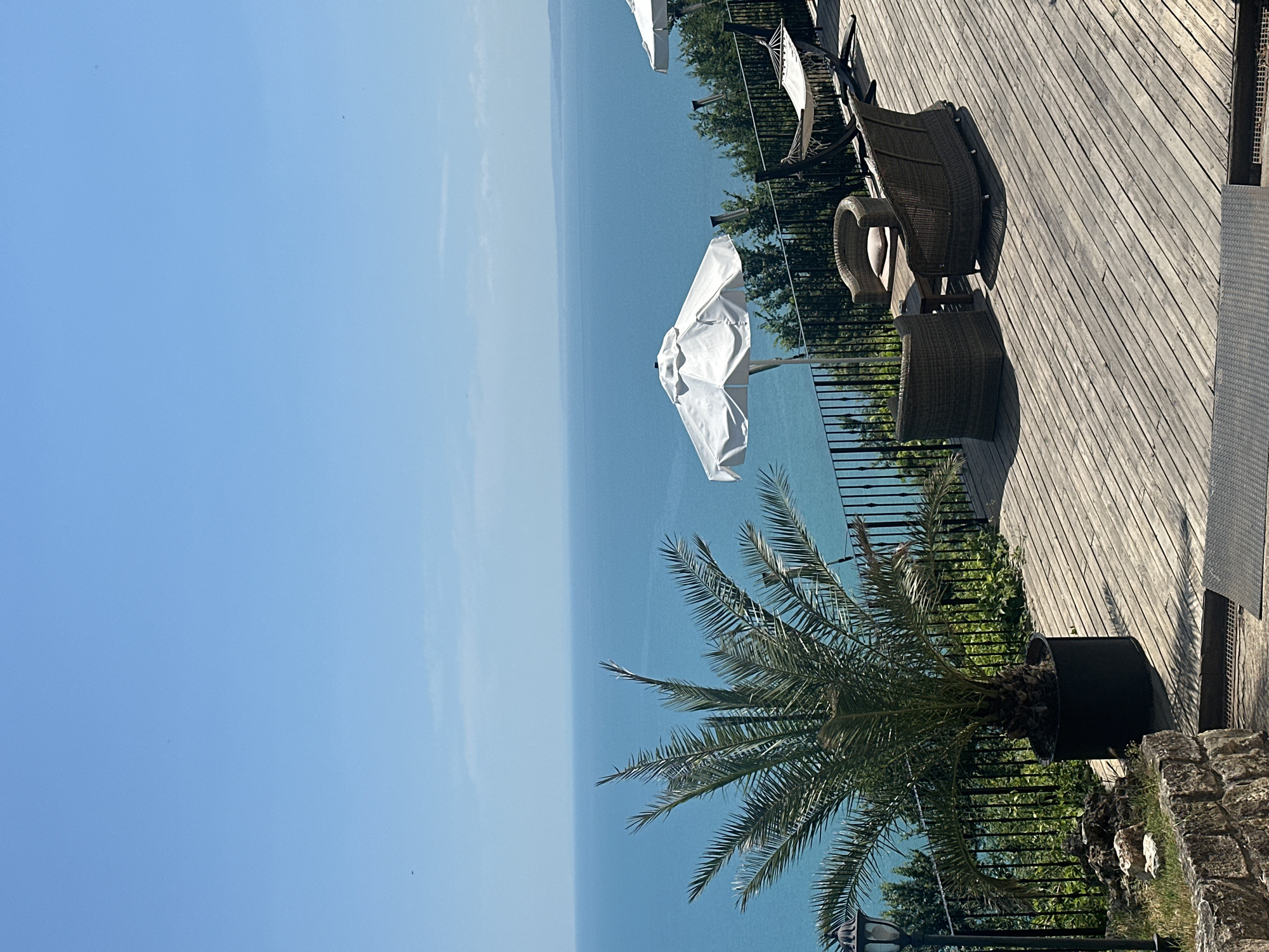 This media file depicts an outdoor scene, likely a terrace or deck overlooking a body of water. The primary subjects are furniture, including several wicker chairs and a matching ottoman, arranged on a wooden deck. A white umbrella, partially open, is also visible, suggesting it is a sunny day and providing shade. In the foreground, a large potted palm tree with lush green fronds adds a tropical element to the setting. A black metal fence separates the deck from a natural landscape with greenery and trees below, leading down to the expansive, calm blue water. The water stretches to the horizon, meeting a clear blue sky with a few wispy clouds. The perspective of the image is a bit tilted, and the lighting suggests it is daytime, possibly late morning or early afternoon, with sunlight casting distinct shadows on the deck. The overall atmosphere appears peaceful and relaxing, an ideal setting for outdoor leisure. There is no visible text in the image.