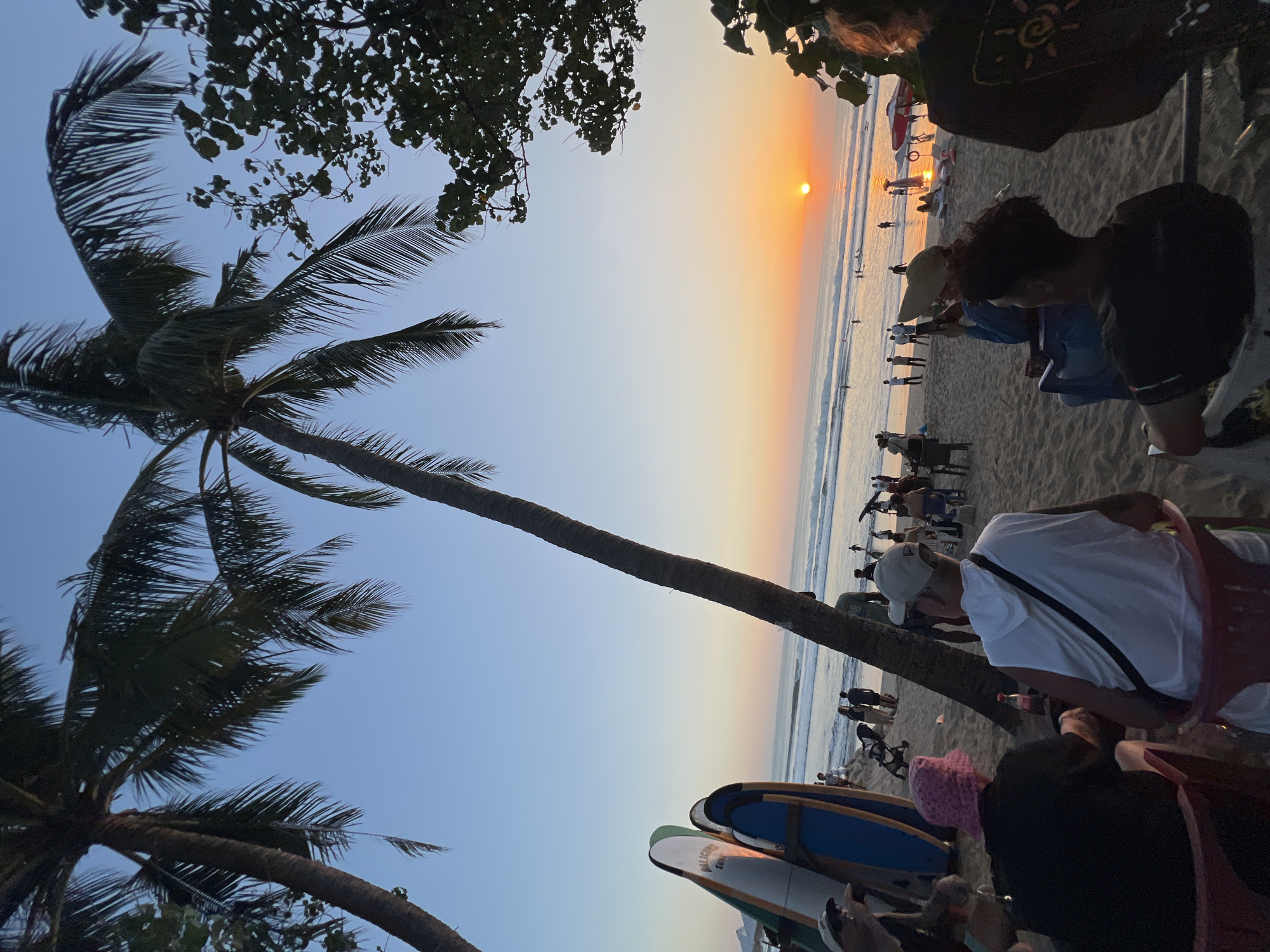 This is a photograph taken at sunset on the beach in Kuta, Indonesia. The sky is a gradient of blue and orange, with the sun just touching the horizon. Palm trees frame the top of the image, their fronds casting shadows. In the foreground, several people are seated on chairs, facing the ocean. Some are engaged in conversation, while others are looking at their phones. In the middle ground, there are more people walking along the beach, and several surfboards are propped up on the sand. The ocean is calm, with small waves rolling in. The scene is peaceful and serene, capturing the beauty of a tropical sunset.