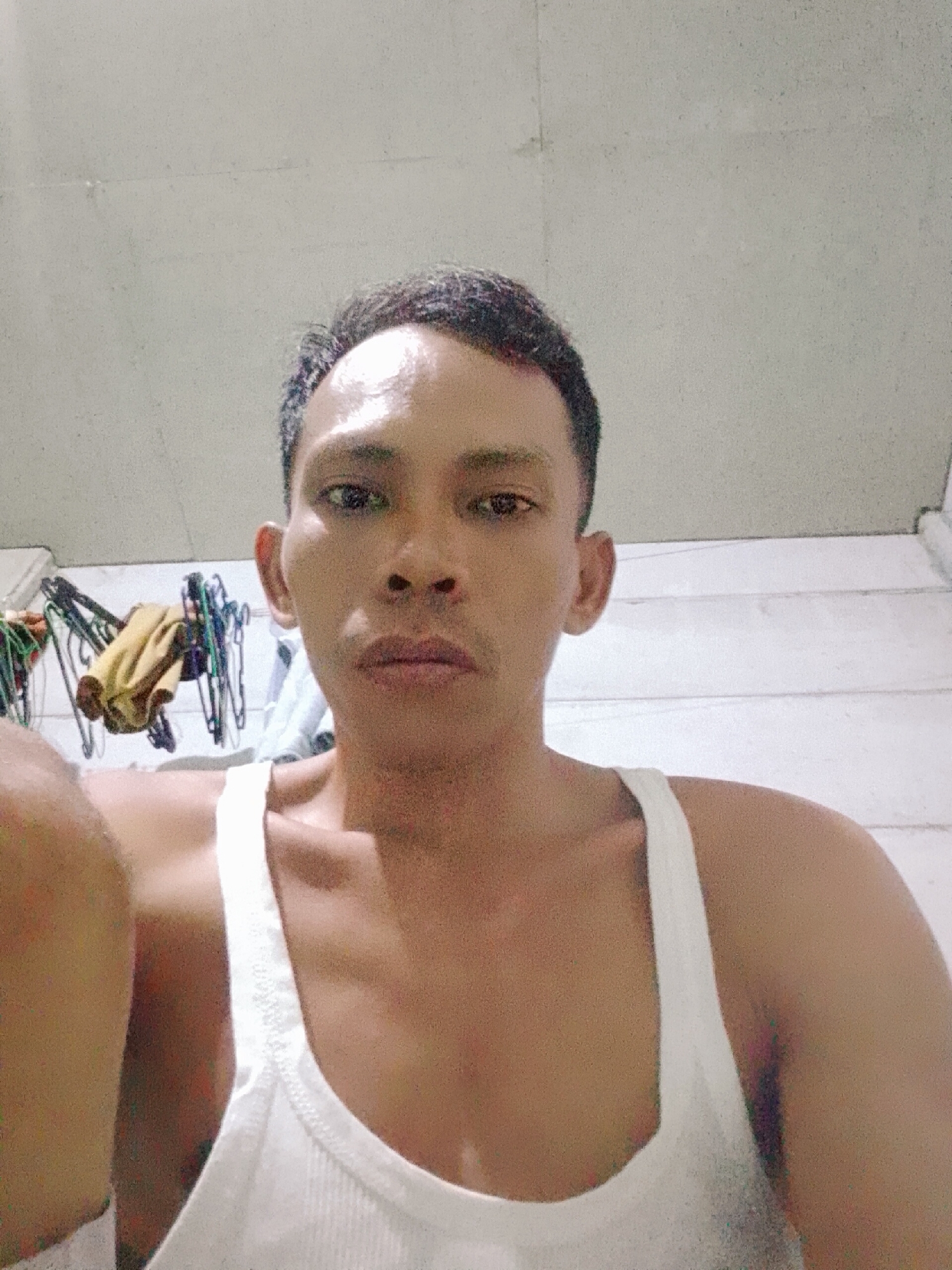 A medium close-up shot shows a man wearing a white ribbed tank top. He is looking at the camera with a neutral expression. The background appears to be a room with a light-colored wall. Clothes hanging on hangers are partially visible in the background. The location is Bukit Ketok, Indonesia.