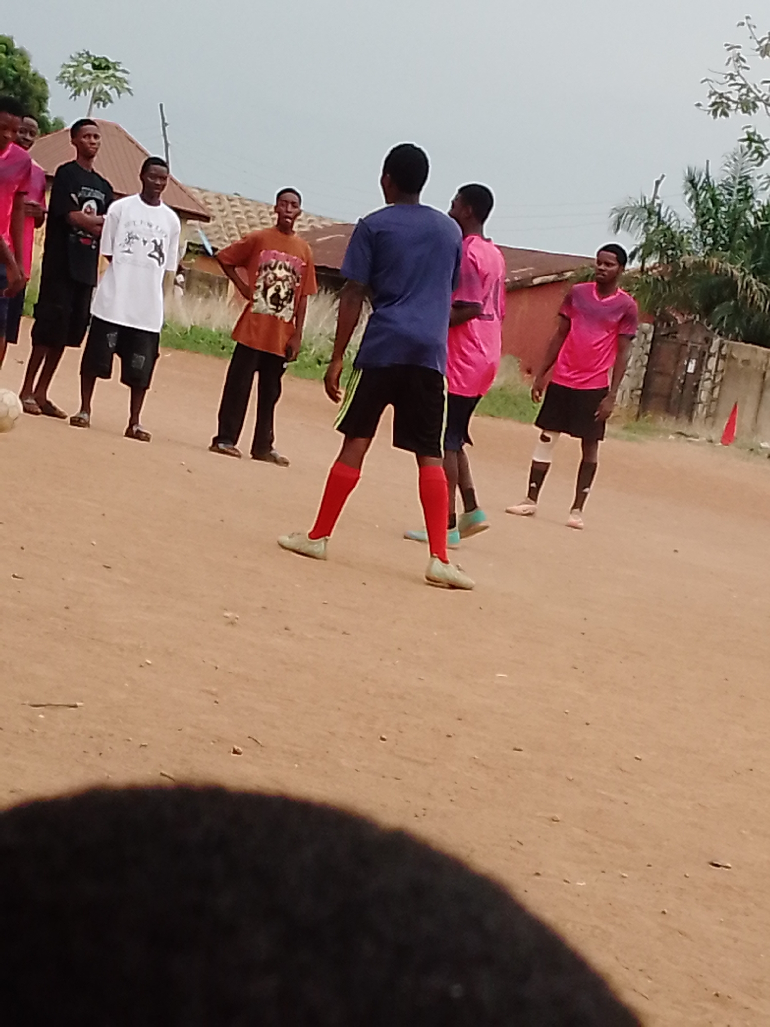 The image shows a group of young men, possibly teenagers, on a dirt field in Unknown, Unknown. They appear to be playing soccer or casually gathered around a soccer ball. The group consists of seven individuals, most of them facing towards the center. They are dressed in various casual outfits, including t-shirts, shorts, and athletic wear. Some of them are wearing sandals or sneakers. The dirt field serves as the playing surface, and in the background, there are buildings and vegetation, indicating a residential area. The sky is overcast.