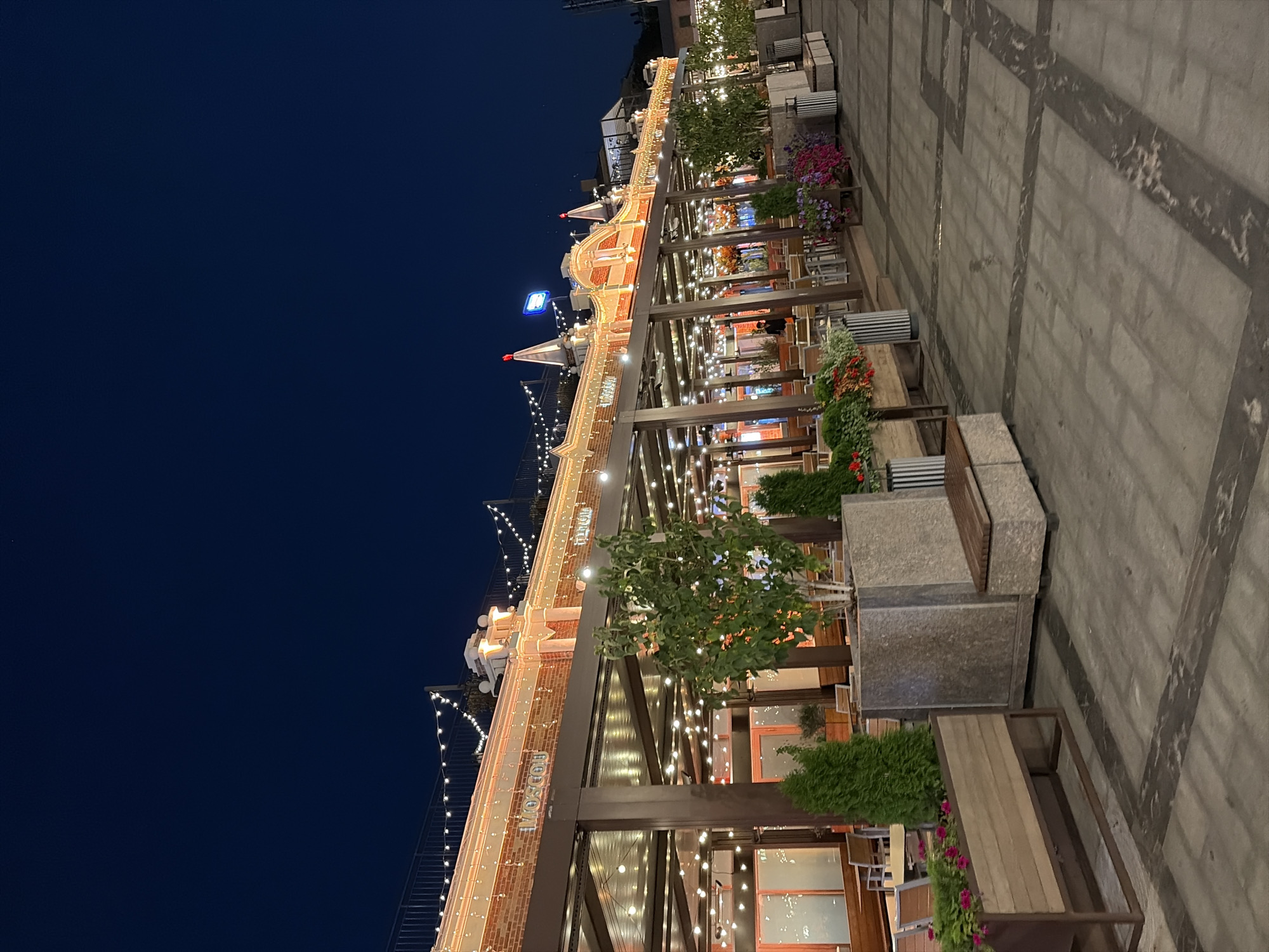 The image depicts an outdoor seating area of a restaurant or cafe in Moscow, Russia, at dusk. The establishment features a long, covered patio illuminated by strings of warm-toned lights. The building facade behind the patio is made of brick and adorned with decorative architectural elements and more string lights. The word "MOSCOW" is visible in raised lettering on the facade.
The patio is furnished with tables and chairs, some of which are visible through the overhang. Several planters containing greenery and flowers are strategically placed along the walkway, adding to the ambiance. The ground is paved with large square tiles, arranged in a pattern with darker rectangular inlays. In the background, a neon sign is visible on a rooftop. The sky is a deep, dark blue, indicating the late hour. No specific actions or interactions of people are clearly discernible.