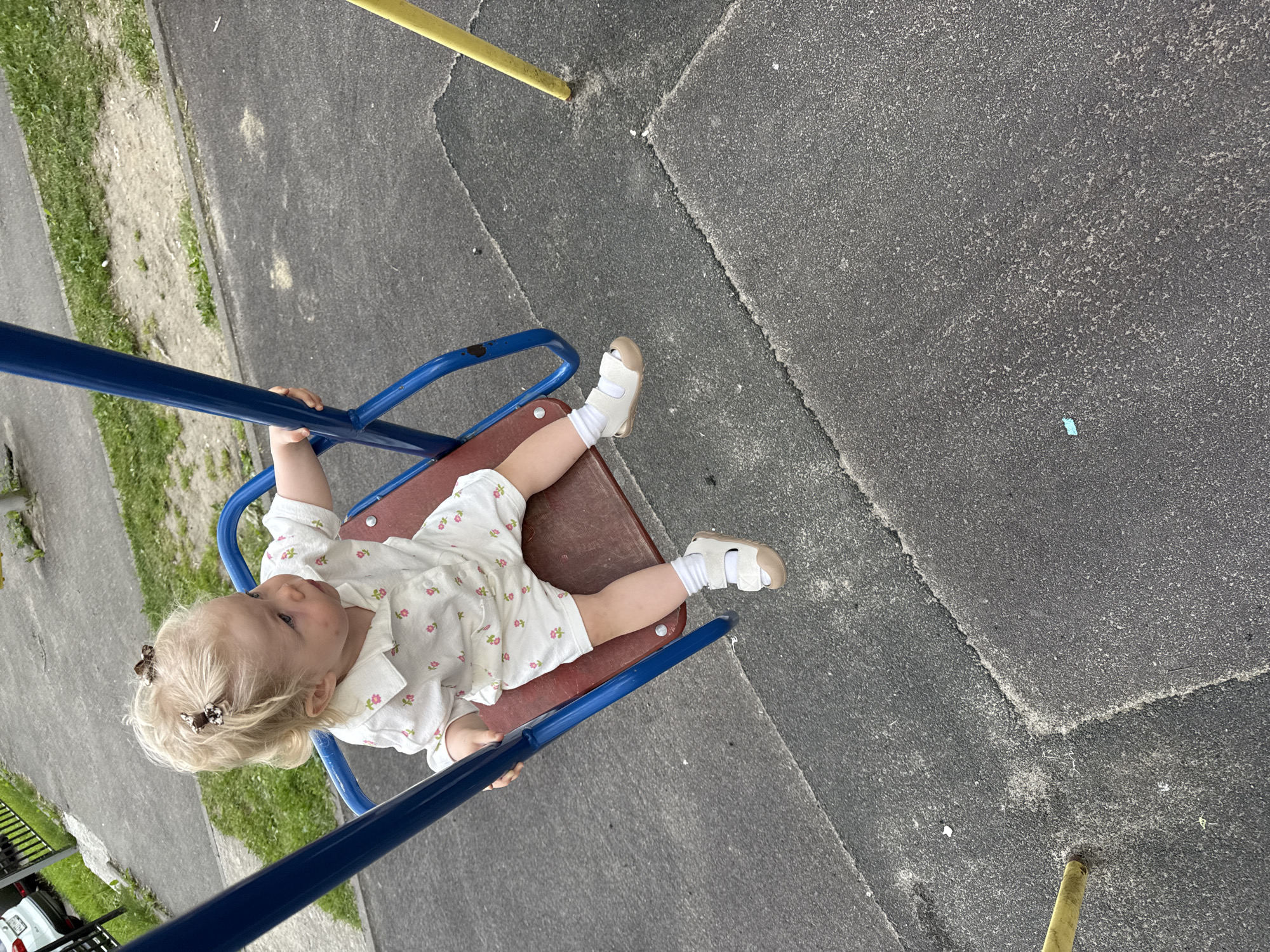 A toddler with blonde hair, adorned with two butterfly-shaped hair clips, sits on a red swing seat attached to a blue swing set frame. The child wears a white short-sleeved shirt with a floral print and matching shorts, white socks, and white sandals. The swing set is located on a paved playground surface composed of dark gray interlocking rubber tiles. In the background, a strip of grass borders a paved walkway, and further back, a black metal fence is visible alongside a parked white car. A yellow pole stands to the right of the swing set. The scene appears to be an outdoor playground in the city of Vladimir, Russia.