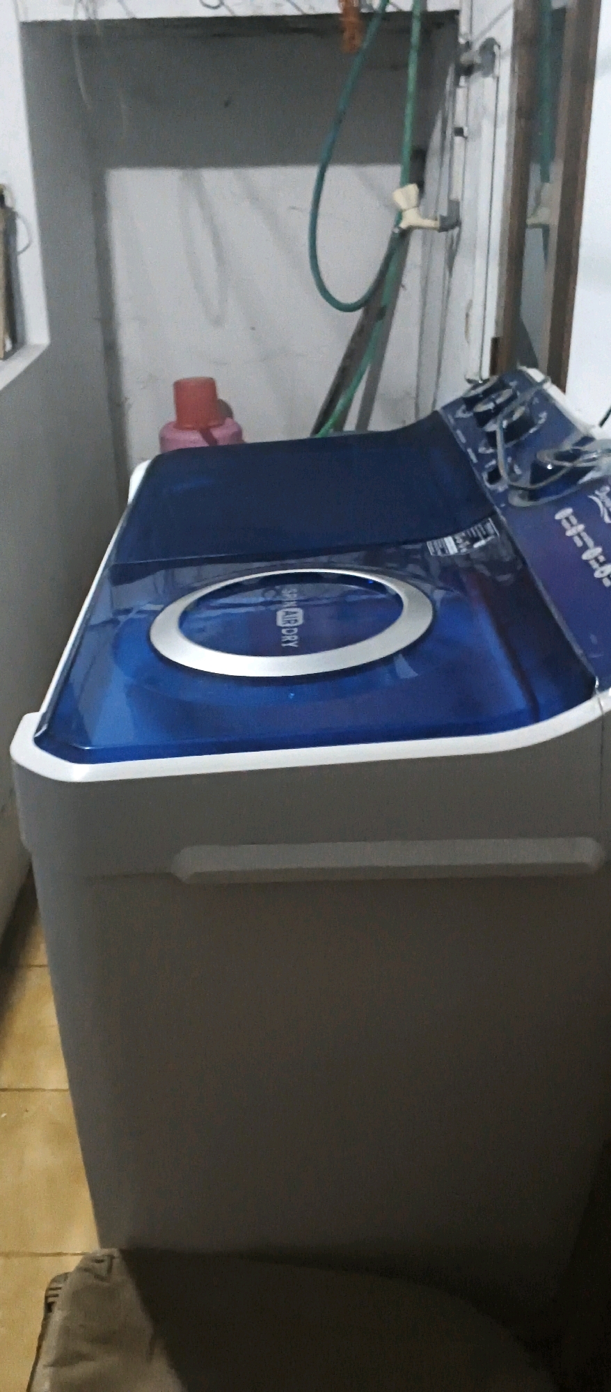 This is a photograph of a top-loading washing machine with a blue lid, located in what appears to be a laundry area or utility room. The washing machine is a dominant subject in the foreground, with its lid closed. The lid is a glossy blue with a silver ring in the center, and text that reads "SPIN AIR DRY" is visible on the lid. The body of the washing machine is a light gray color.
Behind the washing machine, a pale wall is visible, with some plumbing and electrical wiring. A green hose is connected to a faucet on the wall. A red plastic bottle or container is also visible on a shelf or surface behind the washing machine. The flooring appears to be tiled. The overall lighting suggests an indoor environment, possibly a home.
The image appears to have been taken in the city of Cianjur, Indonesia, based on the prompt. However, there are no specific visual cues within the image itself that confirm this location.