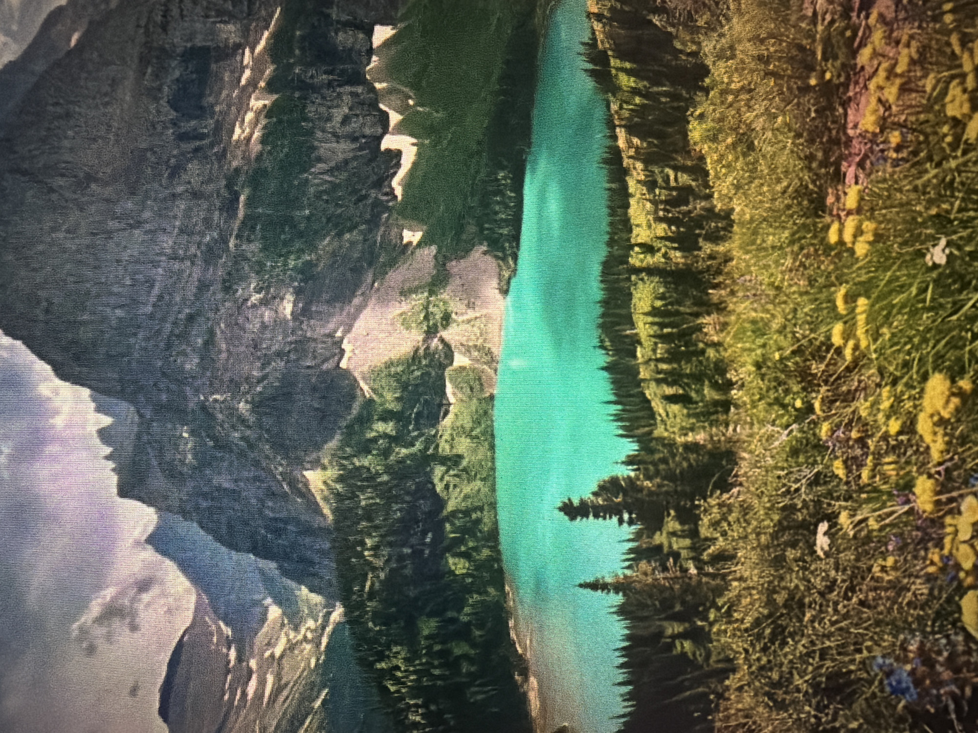 This image depicts a breathtaking mountainous landscape with a vibrant turquoise lake. In the foreground, wild grasses and yellow flowers blanket the ground, leading the eye towards a dense forest of evergreen trees. Beyond the trees, the striking turquoise water of a lake stretches out, reflecting the grandeur of the surrounding mountains. These mountains are rugged and imposing, with dramatic peaks and rocky faces, some of which still show patches of snow. The sky above is a mix of clouds and sunlight, casting shadows and highlights on the terrain. The image does not depict the city of Baan Mae Nam, Thailand; it appears to be a natural landscape, likely in a mountainous region.