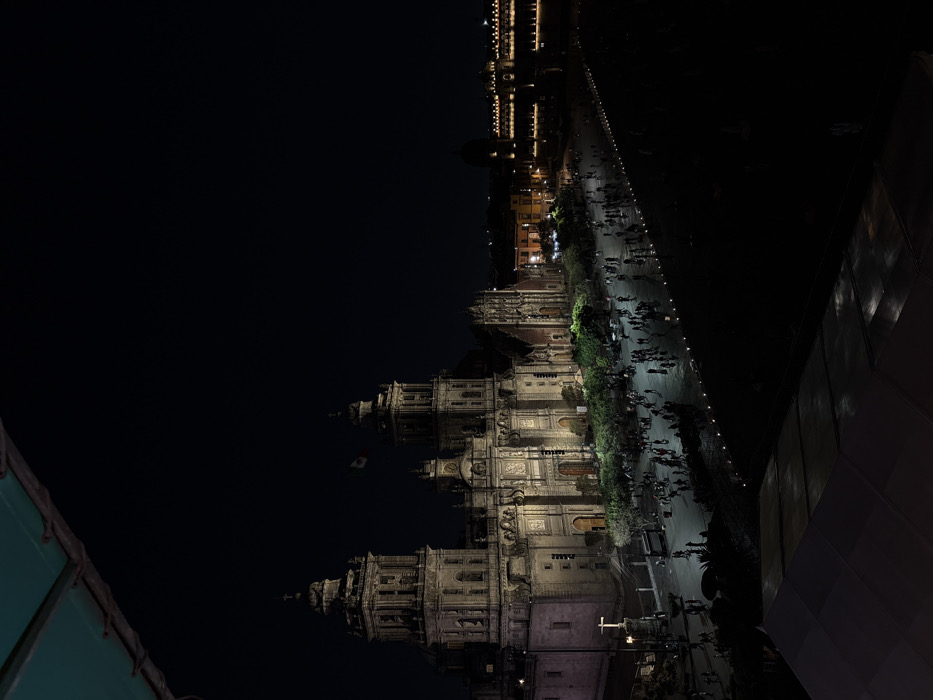 An aerial night view shows the Metropolitan Cathedral of Mexico City. A wide pedestrian walkway filled with people stretches alongside the cathedral, illuminated by streetlights. The National Palace is visible further up the walkway, its lights also reflecting off the pavement. The perspective is slightly tilted, and building edges are partially visible in the corners of the frame.