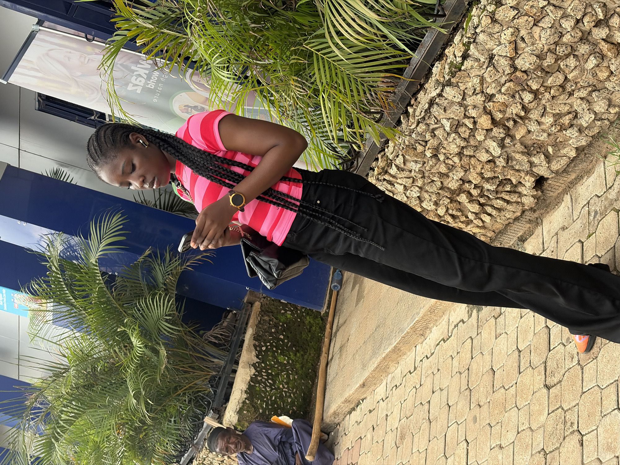 A young Black woman with braided hair, wearing a pink and red striped t-shirt and black pants, stands in front of a stone wall and palm trees. She is looking down at her phone and wearing earbuds and a wristwatch. To her left, an elderly Black man in a purple outfit sits on the ground near a wooden stick. The ground is paved with hexagonal tiles. The scene appears to be outdoors, possibly in Jos, Nigeria, based on the architectural style and vegetation.