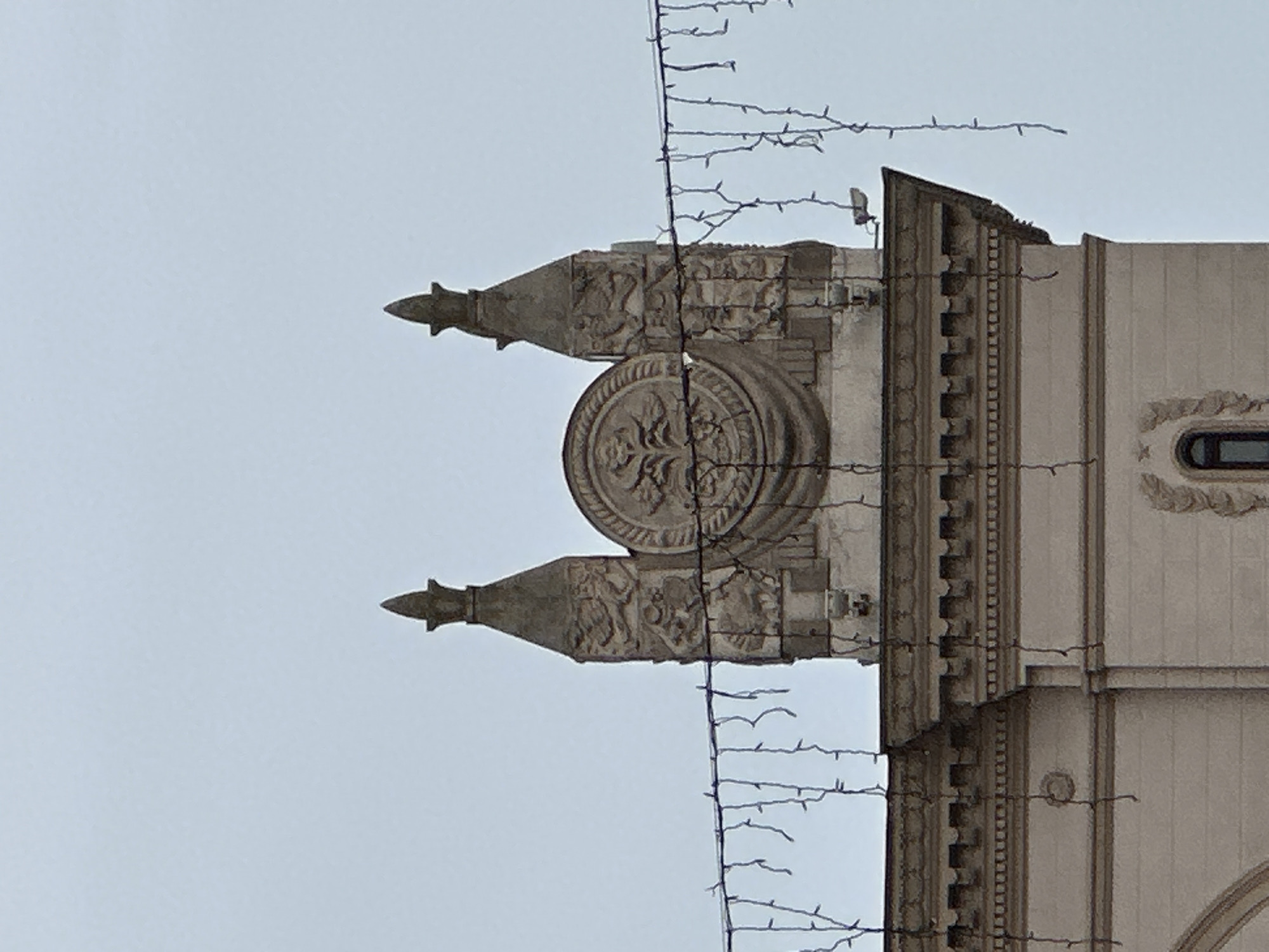 The image displays the upper architectural elements of the Church of Saints Simon and Helena, commonly known as the Red Church, located on Independence Square in Minsk, Belarus, viewed against a light gray, overcast sky. The building material appears to be light-colored stone or stucco. A prominent, ornate cornice line is visible, featuring multiple horizontal bands and a row of dentils. Above this, a central, circular stone medallion in relief sculpture depicts a stylized tree or plant motif. This medallion is flanked symmetrically by two decorative, tapered pilasters or buttresses, each intricately carved with figural reliefs and topped with a pointed finial. Below these elements, on a lower section of the facade, a small, arched window with dark panes is discernible. Multiple strands of decorative string lights, consisting of small bulbs and exposed wiring, are suspended across the mid-foreground, partially obscuring the building's facade. A small, rectangular outdoor floodlight is mounted on the building's upper right edge. No individuals, actions, or interactions are present. The environment consists solely of the architectural structure and the sky.