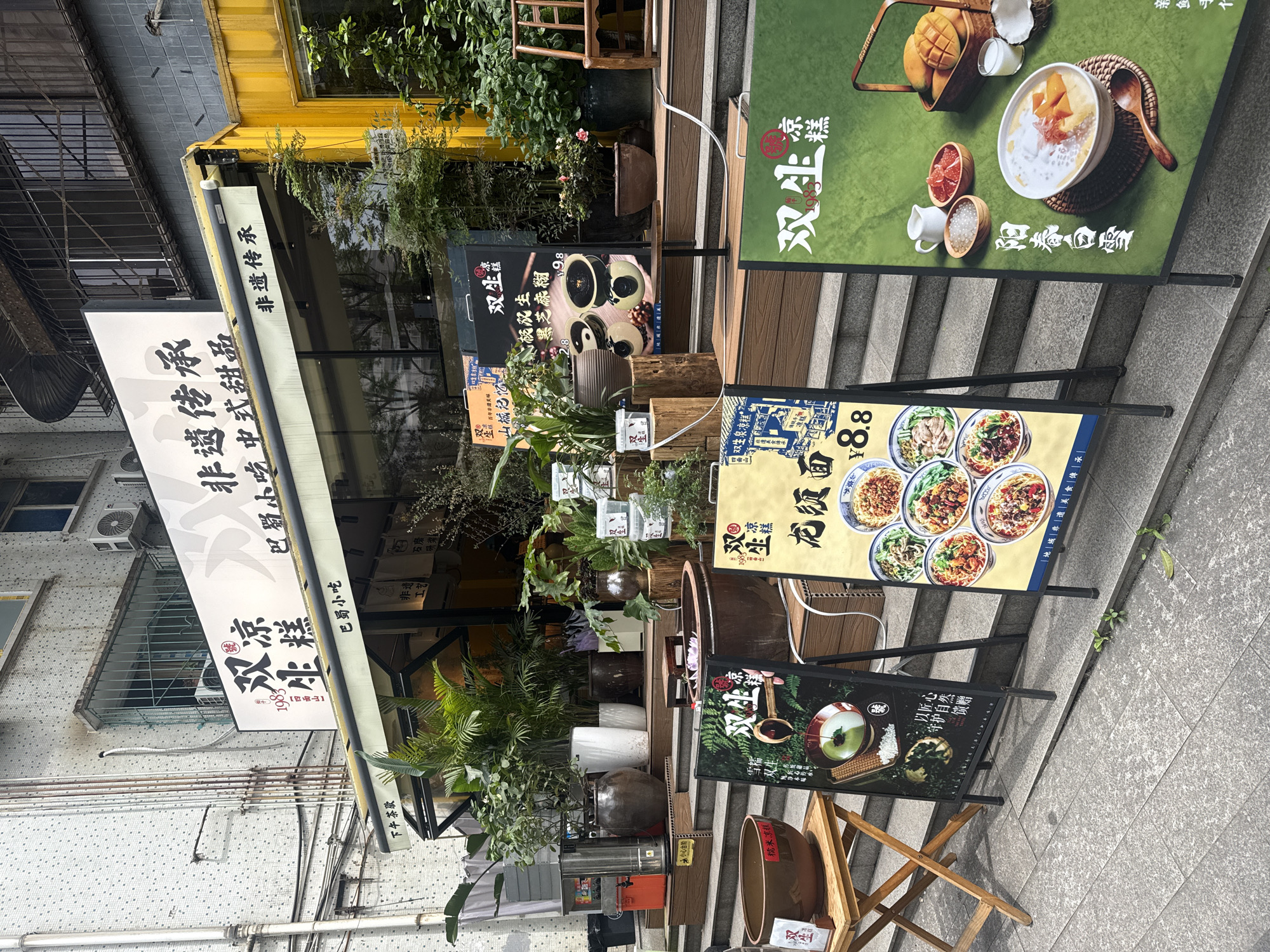 A street-level shot of a storefront in the Yuzhong District of China features a variety of promotional signs and decor. The awning above the entrance has Chinese characters and partially obscures the building's facade. Numerous plants are placed around the entrance. Different menus and advertisements are displayed on stands along the sidewalk in front of the store. The sidewalk is paved with gray stone tiles. The overall scene presents a vibrant and bustling urban environment.