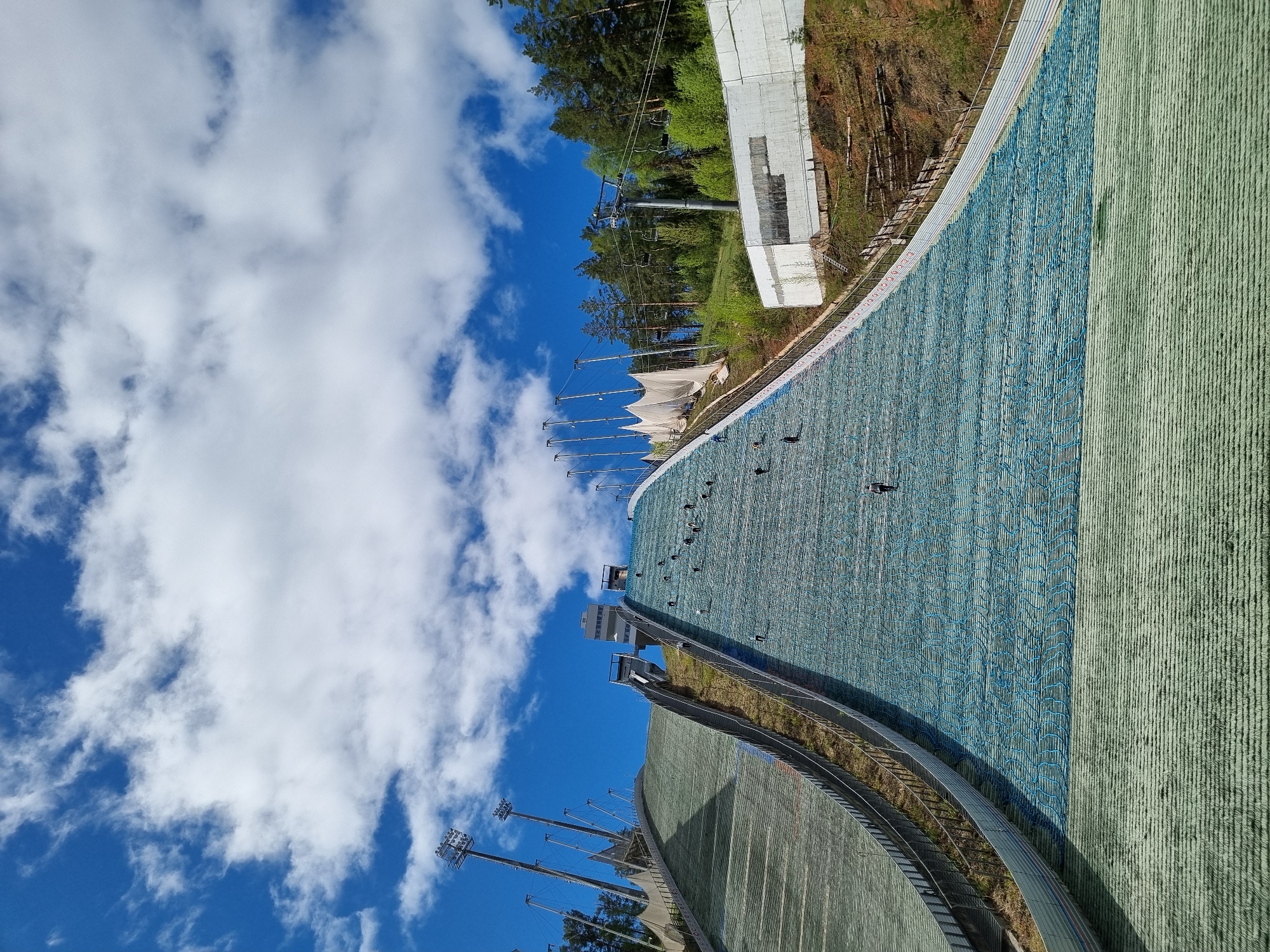The image depicts a ski jumping hill under a partly cloudy blue sky. The slope is covered with artificial turf, transitioning from a green hue to a blue net-like pattern. Visible are the ski jump's inrun, take-off ramp, and landing area. The structure includes supporting towers, floodlights, and possibly a ski lift. In the background, there are trees and a building. The image shows the ski jump's architecture and surroundings. The location is likely the ski jump in Прикамский, Russia.
