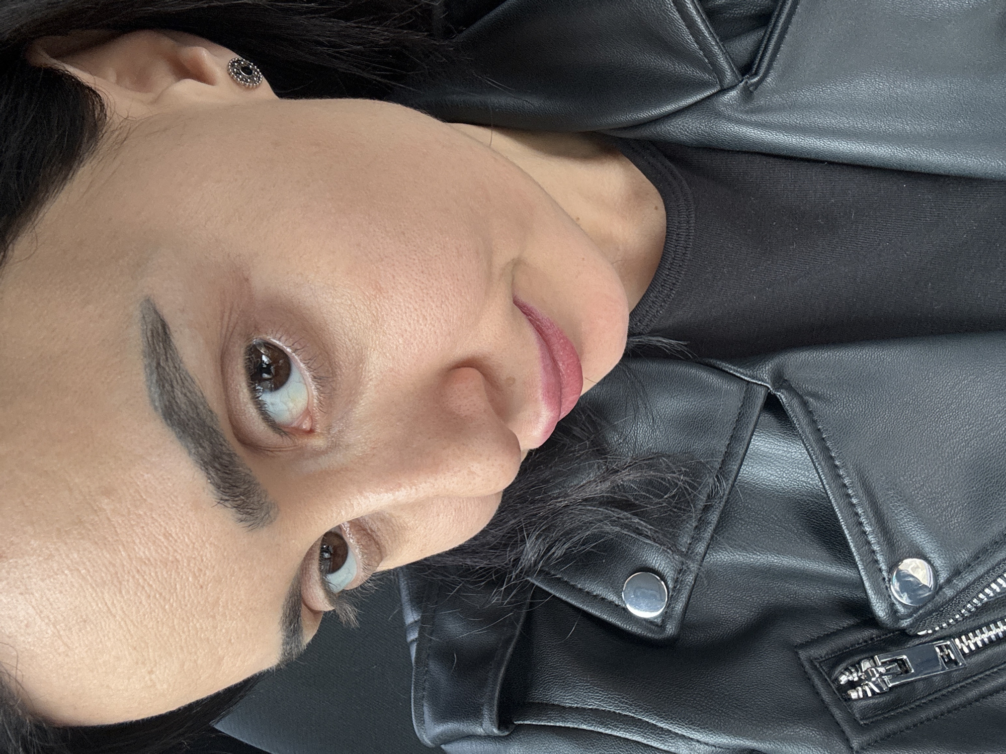 A close-up, rotated image shows a woman with brown eyes and dark hair wearing a black leather jacket and a black shirt. She is wearing a dark-colored earring. No location information can be determined from this image.