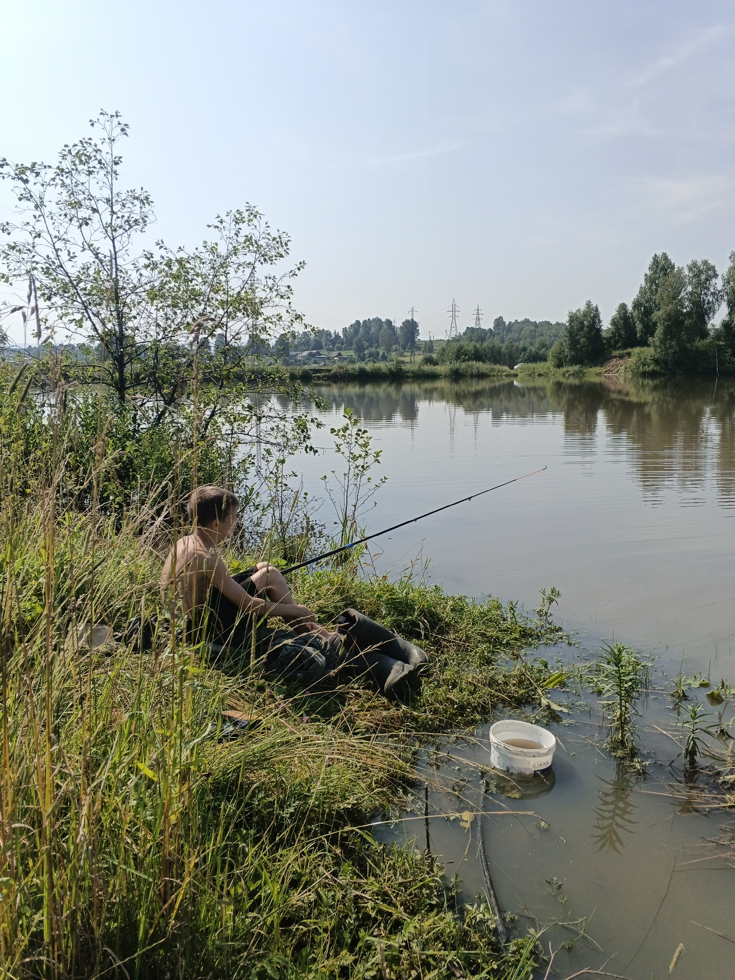 In a rural setting, a young, shirtless man is seen fishing from the bank of a calm body of water. He is sitting on the grassy shore, partially obscured by tall, dry grass, with a fishing rod extended into the water. A white bucket is placed in the shallow water near him. The background features a dense line of trees and a clear sky with a few wispy clouds. The overall scene depicts a peaceful, solitary fishing activity in a natural environment.