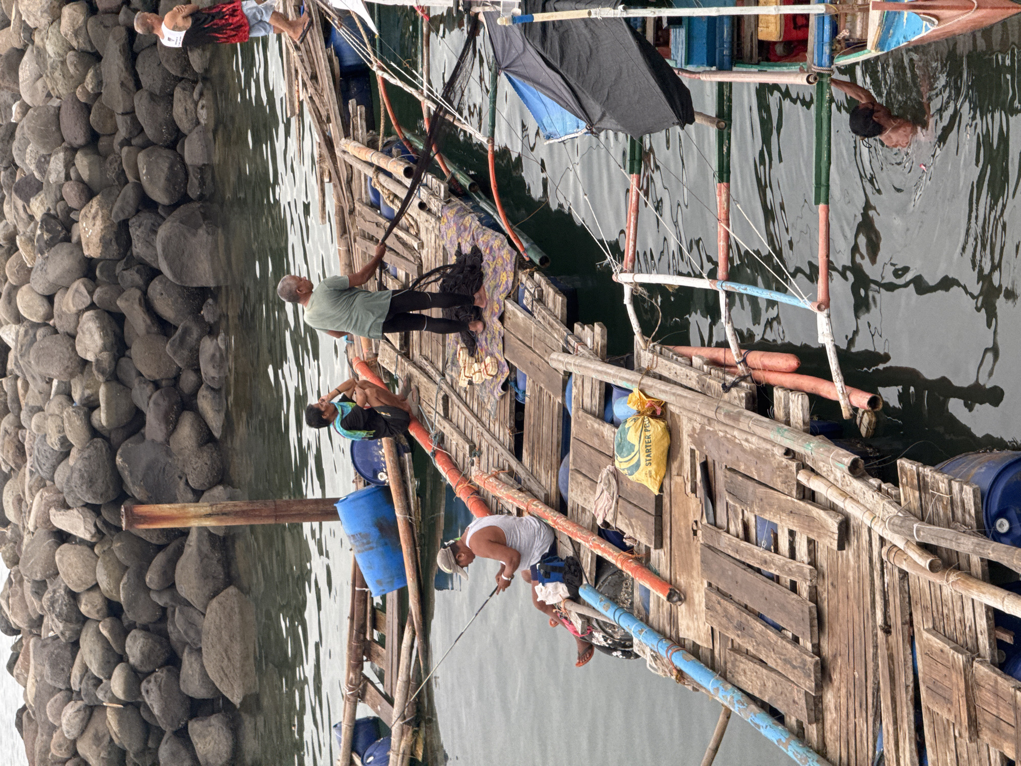 The image shows a group of people on a wooden platform or boat next to a rocky shoreline. One person is swimming in the water next to the platform. Several blue barrels are present, as well as wooden planks and bamboo poles forming the structure. A yellow bag labeled "STARTER PE" is visible. A rocky breakwater or shoreline lies adjacent to the water. The location is likely in Davao City, Philippines. There are 4 people on the platform. One of them is fishing.