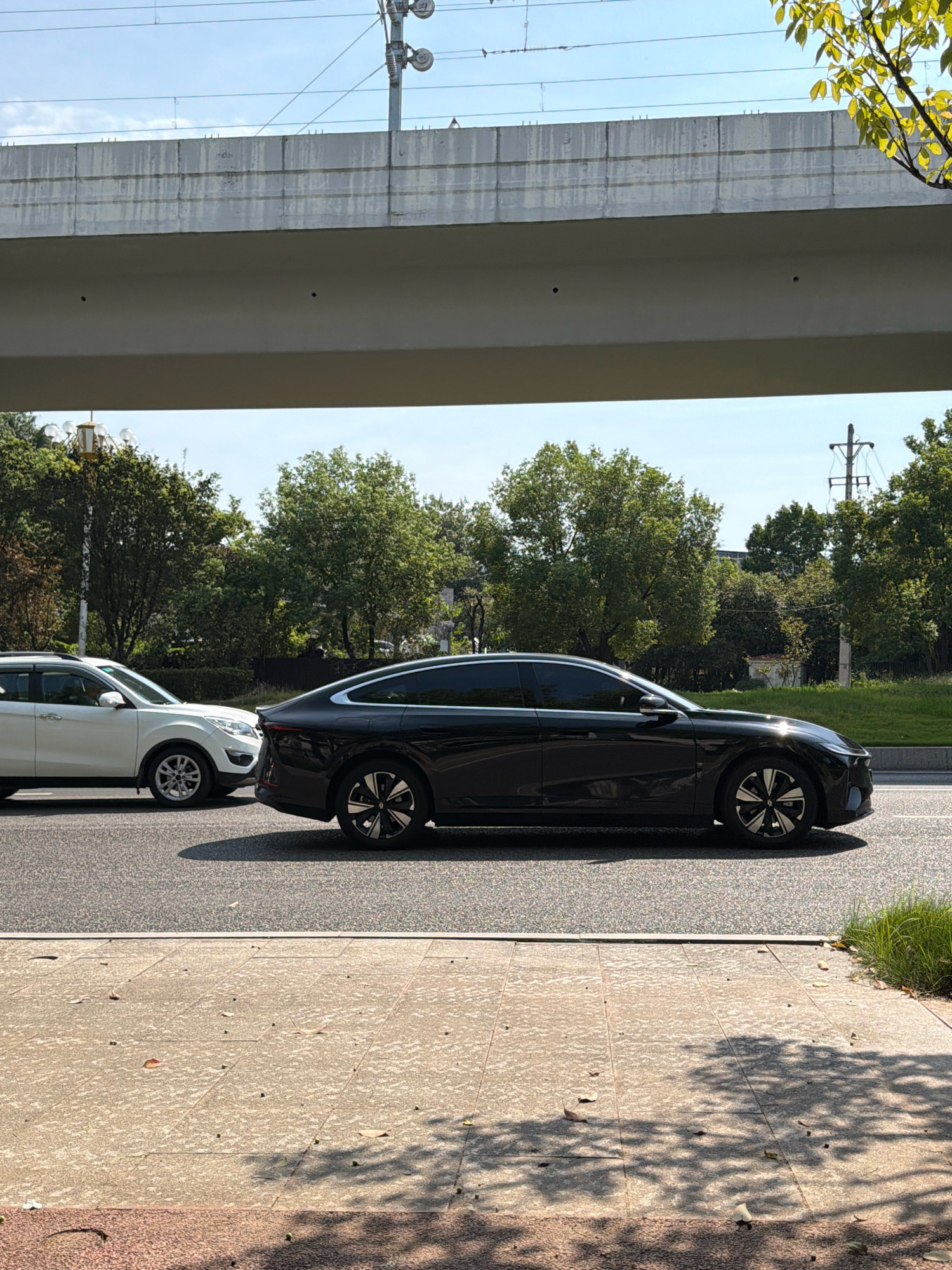 A black sedan and a white SUV are parked on a street in Jiangjin, China, under an elevated structure. The sedan is in the foreground, facing right, with its rear section visible. It is a sleek, modern car with dark tinted windows and distinctive alloy wheels. The white SUV is parked to the left and slightly behind the sedan, also facing right. It appears to be a larger vehicle with visible side mirrors and silver alloy wheels.
The street is paved and lined with a sidewalk. The sidewalk is made of concrete tiles and is scattered with fallen leaves and small green seed pods. Shadows from trees and overhead structures are cast across the sidewalk, indicating a sunny day.
In the background, lush green trees form a dense line along the side of the road. Above the trees, a large, light-colored concrete overpass or bridge spans the scene. Power lines and poles are visible against a clear blue sky with a few wispy clouds. The overall environment suggests an urban or suburban setting in China, likely during daytime.