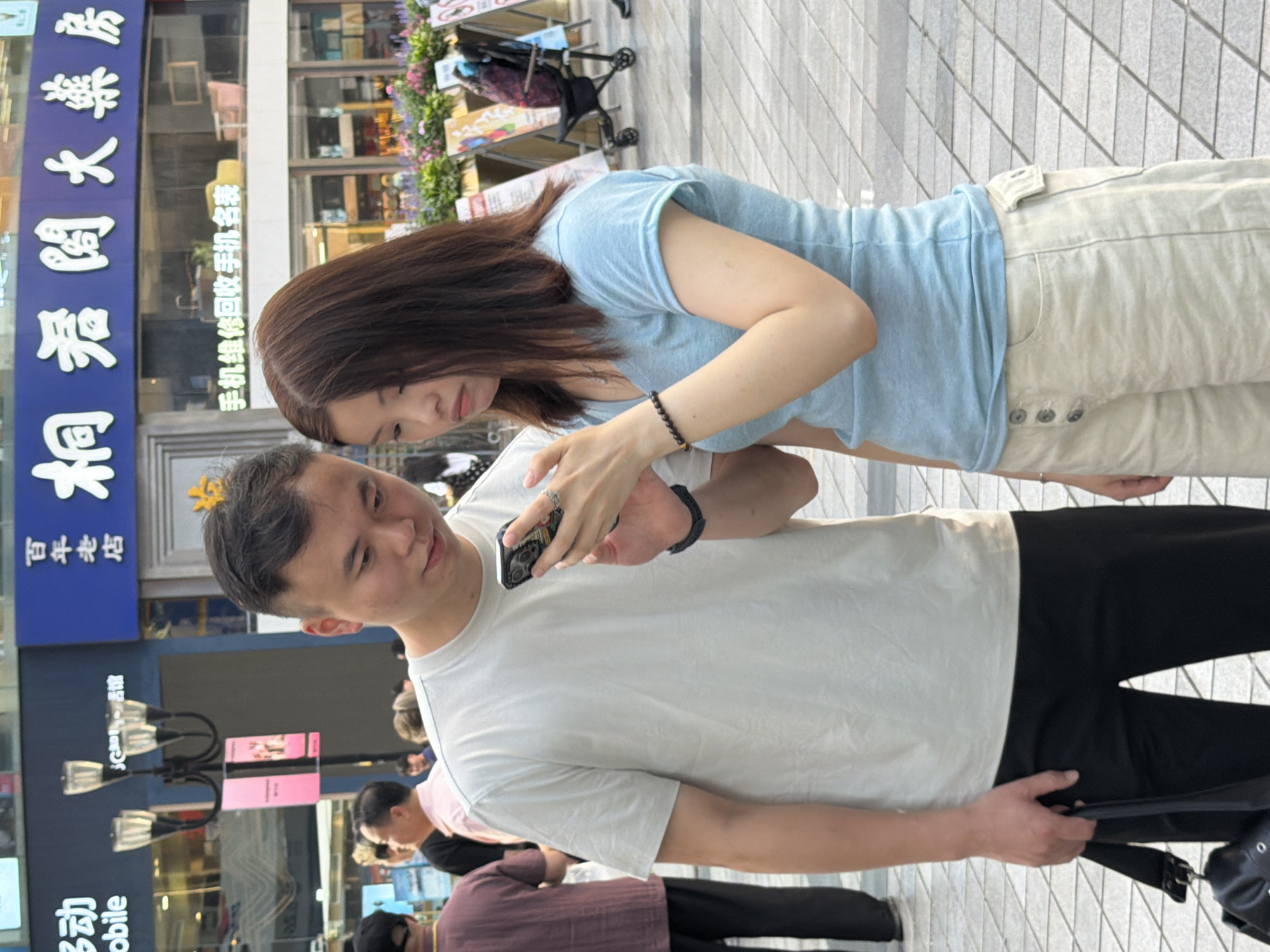 A man and a woman are standing on a city sidewalk in front of a commercial building, looking at a smartphone. The man, on the left, wears a light grey t-shirt and black pants, holding a black bag with his left hand. The woman, on the right, has brown hair and wears a light blue t-shirt and cream-colored shorts. She is holding the phone and the man is looking at it. Behind them, a building displays a large sign with Chinese characters and a smaller sign that reads "手机维修回收手机名表" (mobile phone repair, recycling, watch sales). The sidewalk is paved with grey square tiles, and in the background, there are flower displays and other pedestrians. The scene appears to be in Yuzhong District, China.