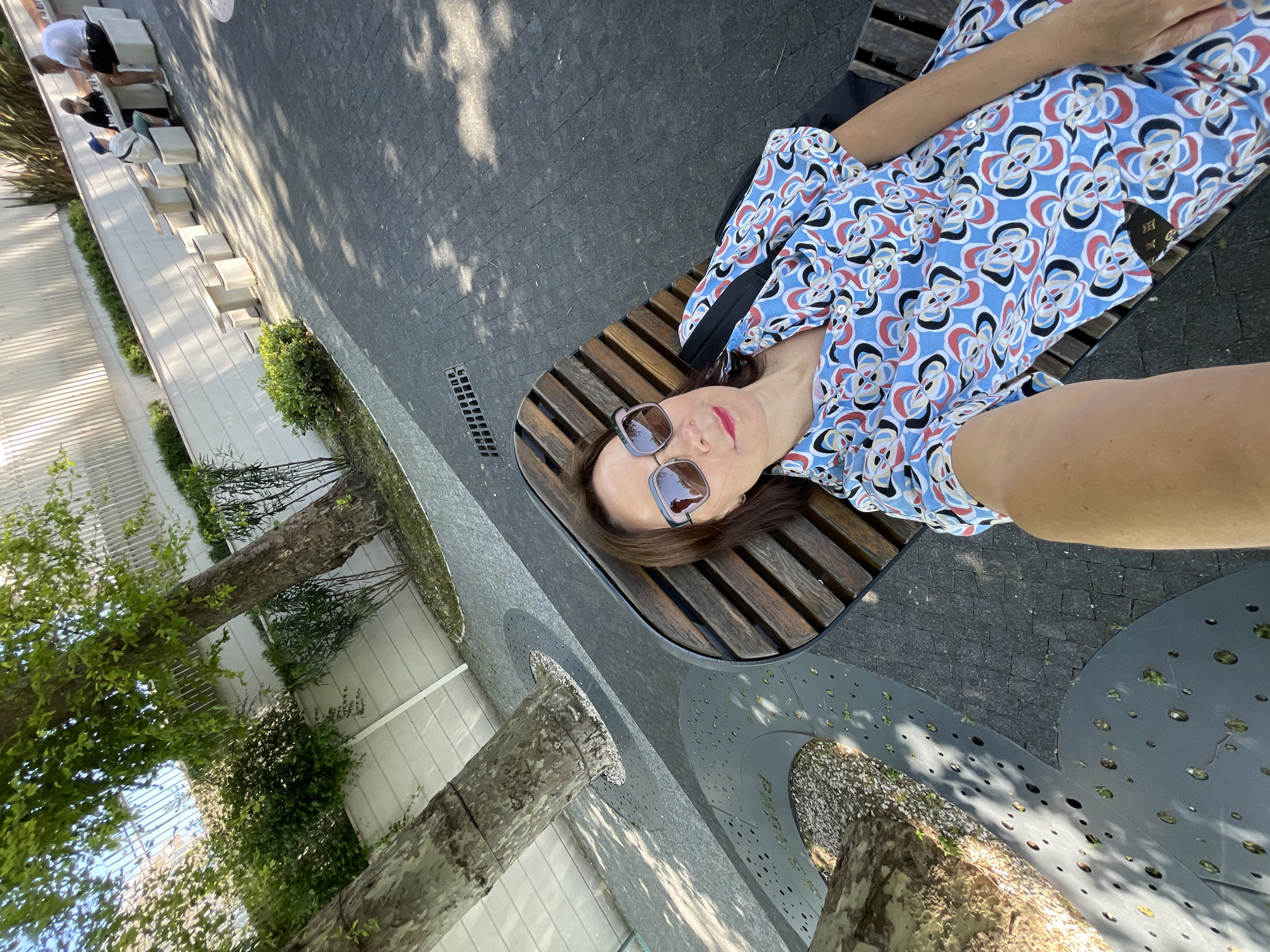 A woman is captured in a selfie, lying on her back on a slatted wooden bench in an outdoor park setting. She wears a blue dress with a prominent pattern of red, black, and white abstract shapes, and large sunglasses. A relaxed expression is visible on her face. Beside her, a portion of a bag, possibly with a Louis Vuitton monogram, is seen.
The bench is situated on dark grey paved ground, likely cobblestones or tiles, dappled with sunlight and tree shadows. In the upper left background, two men and a woman are seated on contemporary light-colored stone benches. Further back, a white wall or building is partially obscured by green shrubs. Several trees with light-colored bark punctuate the paved area. Around the base of some trees are modern, circular, perforated metal grates, one of which clearly displays the Cyrillic word "ПАРК" (PARK).
The scene is brightly illuminated by strong daylight, indicating a sunny midday or early afternoon. The overall environment suggests a modern, well-maintained public park or plaza in Sochi, Russia. The image is oriented 90 degrees clockwise from standard upright viewing.