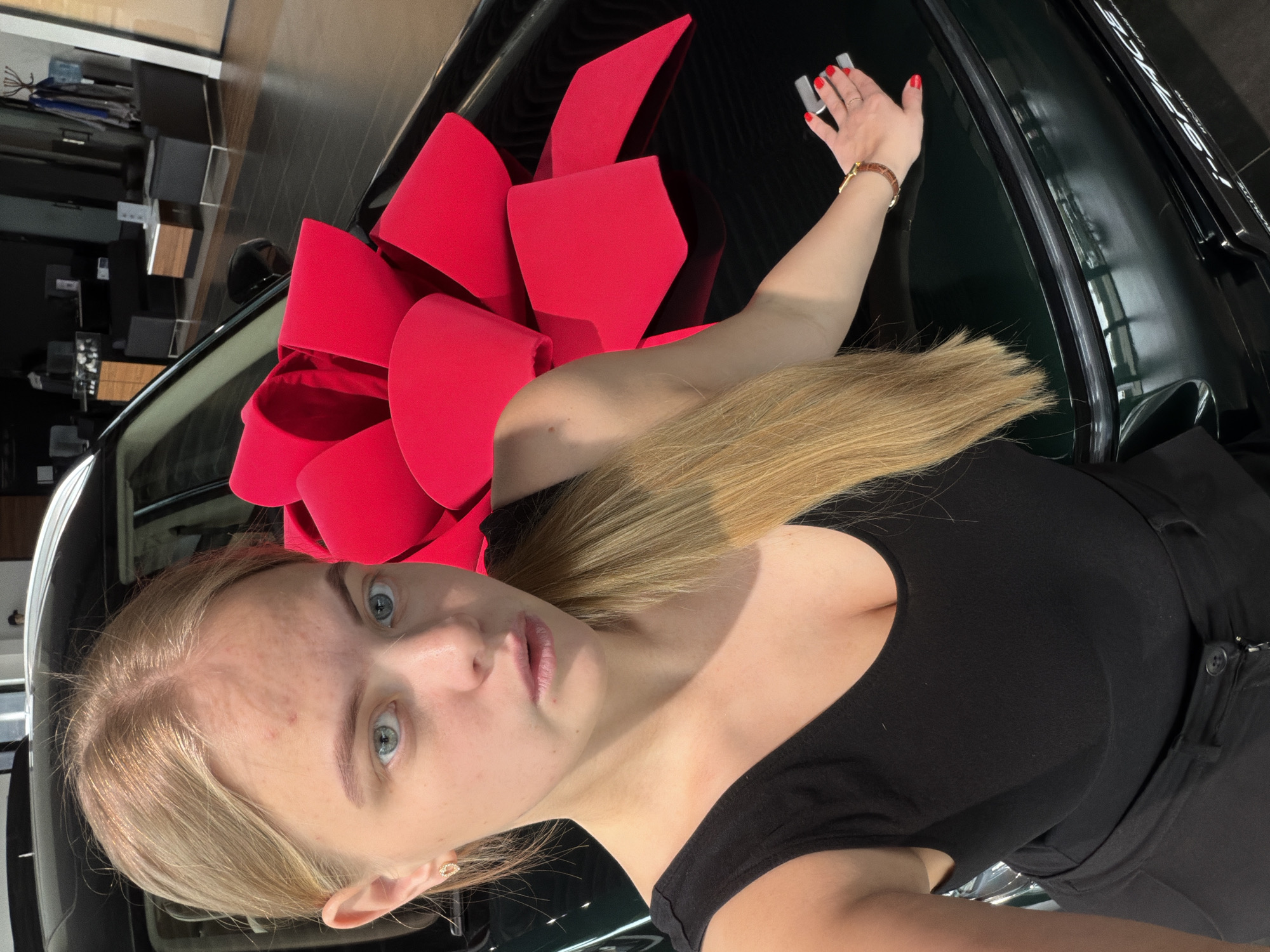 A blonde woman in a black top poses for a selfie next to a green car with a large red bow on the hood. The woman has fair skin, blue eyes, and is wearing a watch on her left wrist. Her left hand is extended towards the windshield. The background shows a car dealership or showroom with visible boxes and reflective surfaces. No location details of Izhevsk, Russia are discernible.