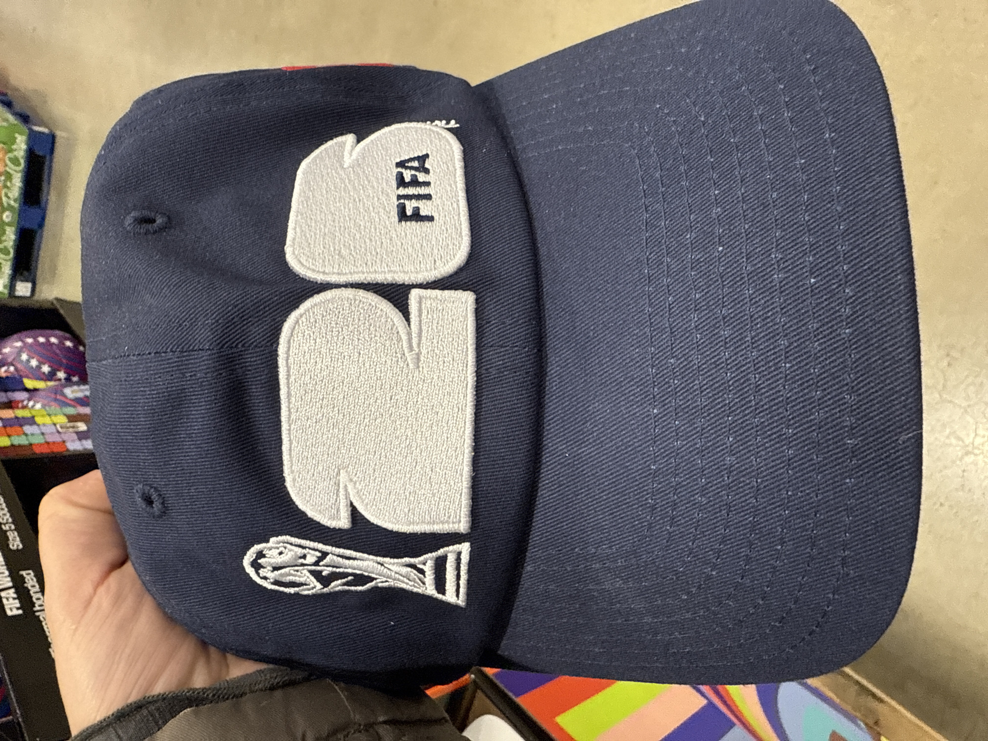 A person is holding a dark blue baseball cap with white embroidered designs in a retail environment. The embroidery includes "FIFA" stacked vertically above the number "26" and a stylized trophy at the bottom. The hat appears to be new and is being held over a display of packaged goods with colorful designs. The location is Bolingbrook, United States of America, as determined by the retail items in the background.