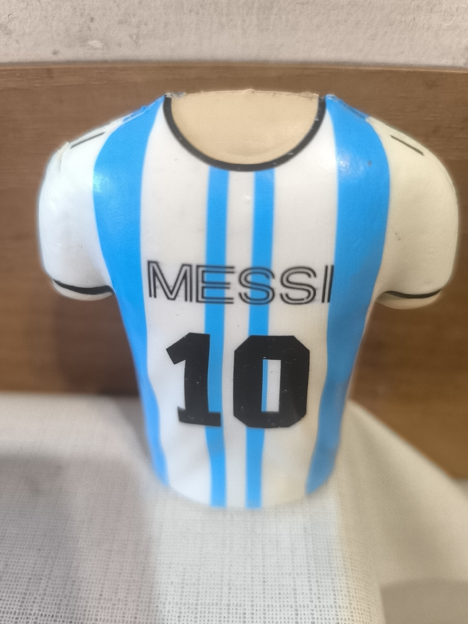 A close-up, eye-level shot in Yerevan, Armenia, captures a miniature replica of a football (soccer) jersey, likely a decorative item such as a cake topper or collectible figurine. The jersey is predominantly white with five vertical light blue stripes on the back. A thin black line outlines the collar and the cuffs of the short sleeves.
Prominently displayed on the back of the jersey is the name "MESSI" in black, hollow block letters. Below the name, the number "10" is printed in solid black, signifying the iconic number worn by Lionel Messi for the Argentina national team. The object appears to be made of a rigid material, possibly plastic or ceramic, with a slight sheen. There are minor imperfections and small chips visible on the edges of the collar and sleeves, suggesting its manufacturing process or handling.
The jersey rests on a white, textured fabric surface, which shows a subtle grid pattern. In the background, a warm-toned wooden surface, possibly a table or shelf, is visible, adding depth to the scene. No activity is taking place; the image is a static portrayal of the decorative item.