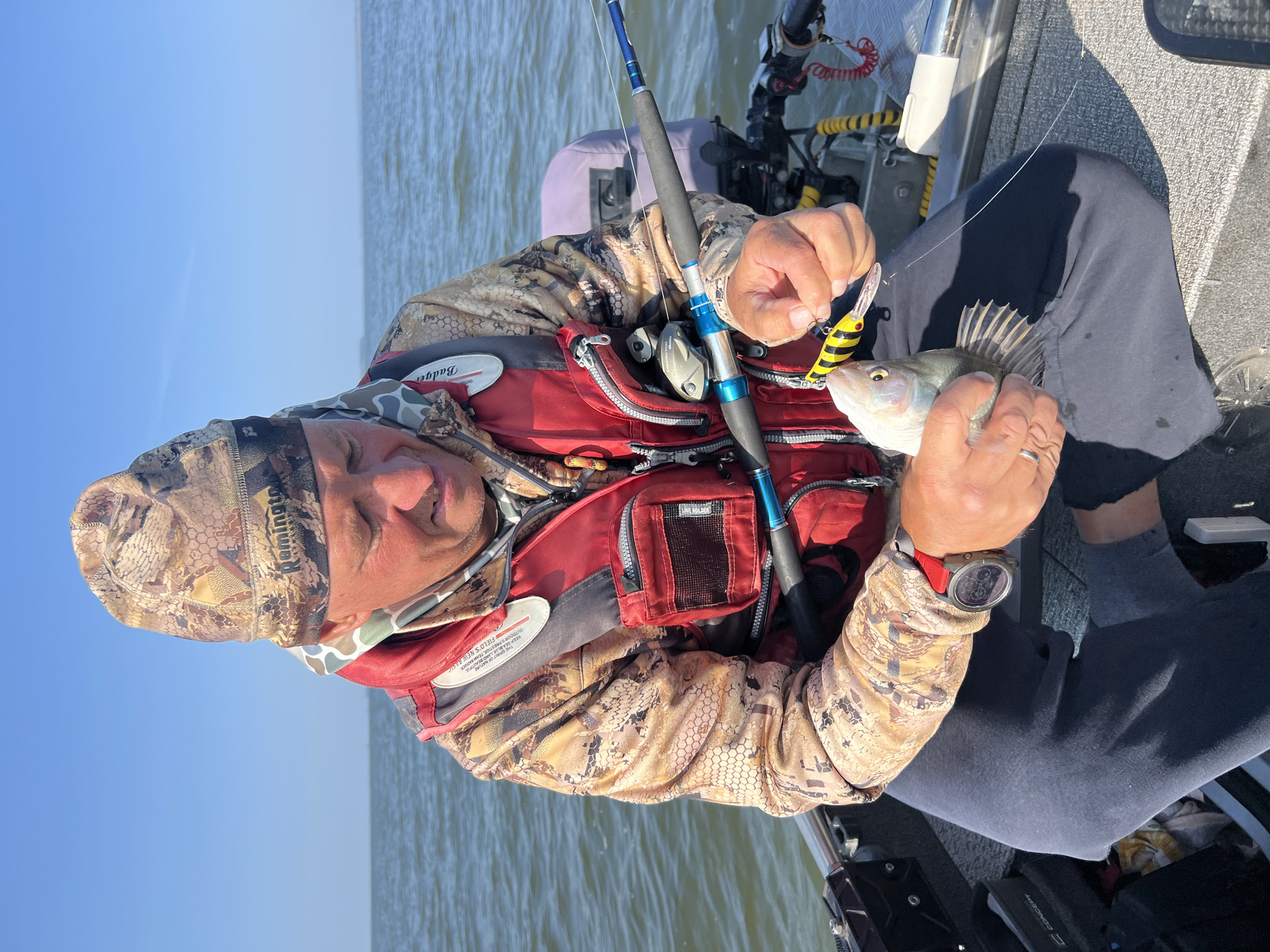 A man wearing a camouflage beanie, camouflage jacket, and a red life vest is sitting in a boat on the water. He is holding a small fish in his left hand and a fishing lure in his right hand. The fishing rod is visible next to him. The sky is blue and clear. The environment appears to be a lake or a large body of water. The man is looking down at the fish and lure.