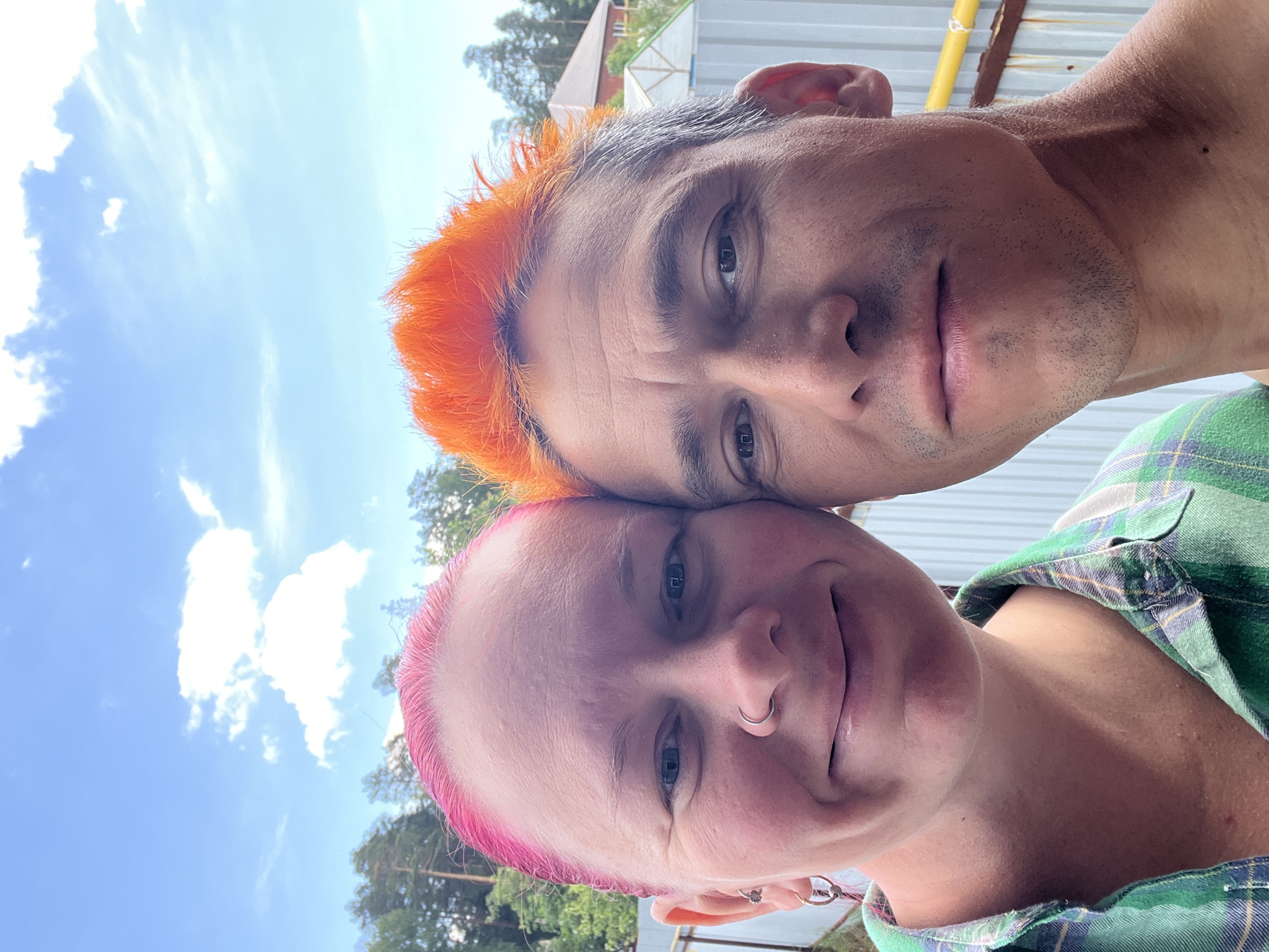 This image is a close-up outdoor selfie taken in the city of Chaikovsky, Russia, featuring two adults positioned closely together. On the left is an individual with short, bright pink hair, blue eyes, a hoop nose piercing in their left nostril, and multiple ear piercings in their left ear. This individual is smiling and wears a green and blue plaid collared shirt. On the right is an individual with short, spiky, bright orange hair on the top of their head, transitioning to gray hair on the sides, with dark eyes and light facial stubble. Their expression is neutral to faintly smiling, and their right shoulder and neck are visible, suggesting they are shirtless or wearing a low-cut garment. Their heads are nearly touching in the center, and both are looking directly at the camera. The background features a clear blue sky with scattered white clouds, dense green coniferous trees, a distant house with a brown pitched roof, and a light-colored corrugated metal fence, with a horizontal yellow pipe visible behind the male subject. The scene is illuminated by strong natural light.