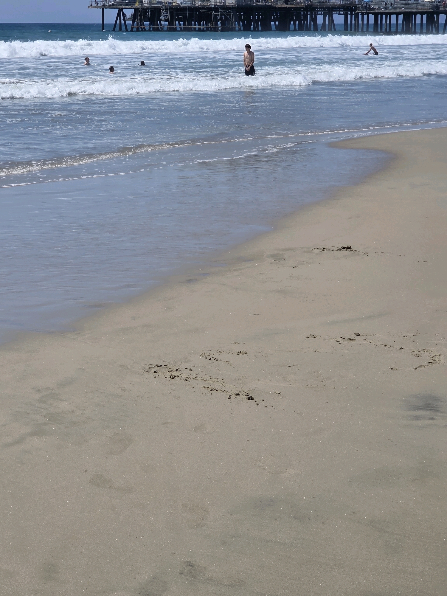 A view from a sandy beach shows a pier in the background, with several people in the ocean. The pier has multiple levels and appears to be made of wood and metal. The water is blue and has white waves breaking near the shore. One person is standing in the water on the left, another is sitting in the water in the middle, and two more are swimming further out. The beach is mostly empty, with some footprints in the sand. The sky is blue and clear. This scene is likely the Santa Monica Pier in Santa Monica, California.