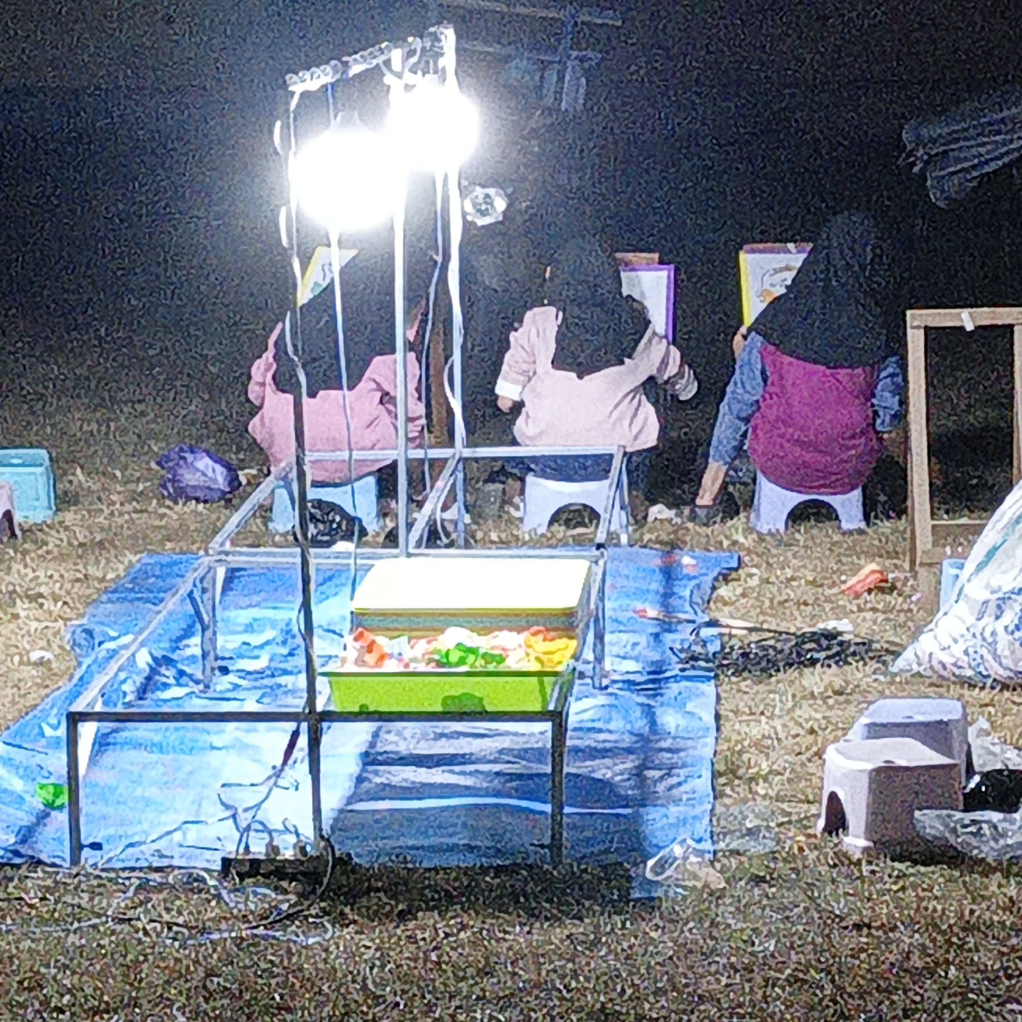 The image depicts an outdoor scene at night in Tanahbaya, Indonesia, illuminated by a bright overhead light. Three individuals, all appearing to be women wearing hijabs, are seated. One person in the foreground is wearing a pink jacket, another in the middle ground is in a light-colored jacket, and the third person on the right is wearing a dark hijab and a purple vest over a blue shirt. They appear to be engaged in some activity, possibly drawing or crafting, as there are easels and art supplies visible. In the foreground, a metal frame structure supports a tray filled with colorful objects, possibly toys or craft materials, which is covered by a blue tarp. The ground is covered in dry grass and debris. Several small, stool-like objects are scattered around the scene, along with some bags and plastic items. The overall atmosphere suggests a nighttime event or gathering.