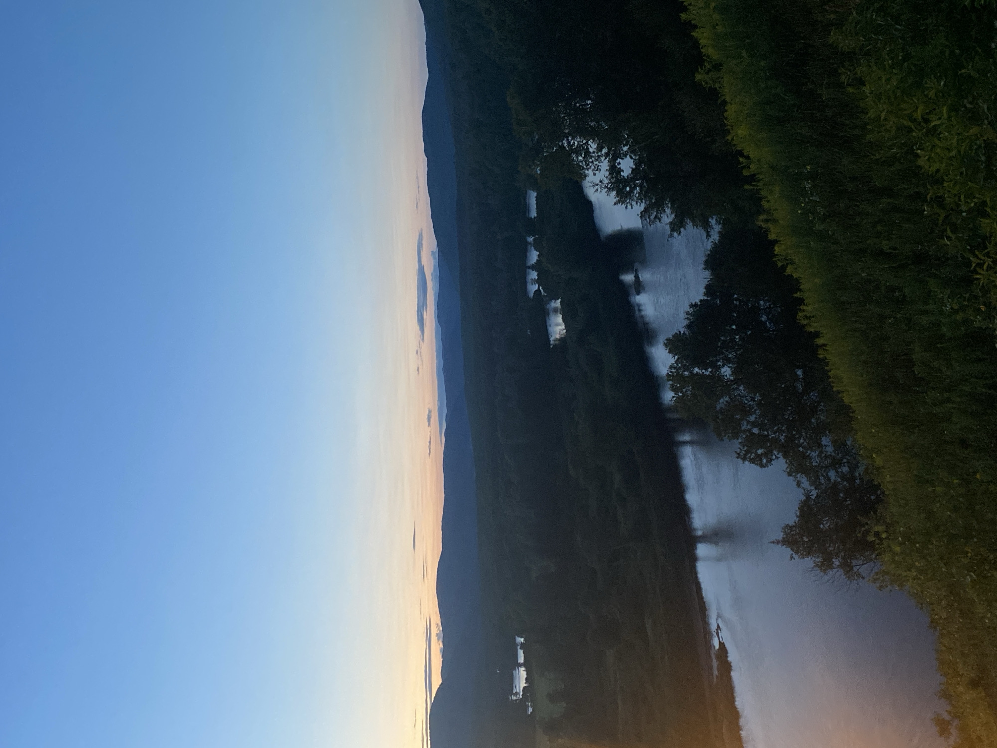 This is a high-angle, wide shot of a river valley at dusk in Saint-Gabriel-de-Valcartier, Canada. The sky is a clear, pale blue, transitioning to a faint orange and pink glow near the horizon. Silhouetted against the sky are rolling hills covered in dark green forests. A wide, calm river flows through the valley, reflecting the dim light. The foreground is dominated by a steep, grassy embankment covered in lush green vegetation and some yellow wildflowers. The overall scene conveys a peaceful, natural landscape at the end of the day.